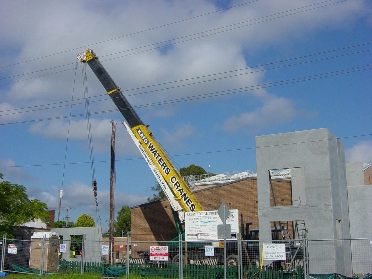 Construction site with a crane labeled 'KATO WATERS CRANES' lifting materials near a partially built concrete structure under a blue sky with clouds.