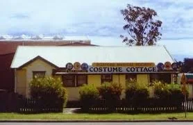 A building with a sign that reads 'Costume Cottage' and several pumpkins on the roof, surrounded by greenery.