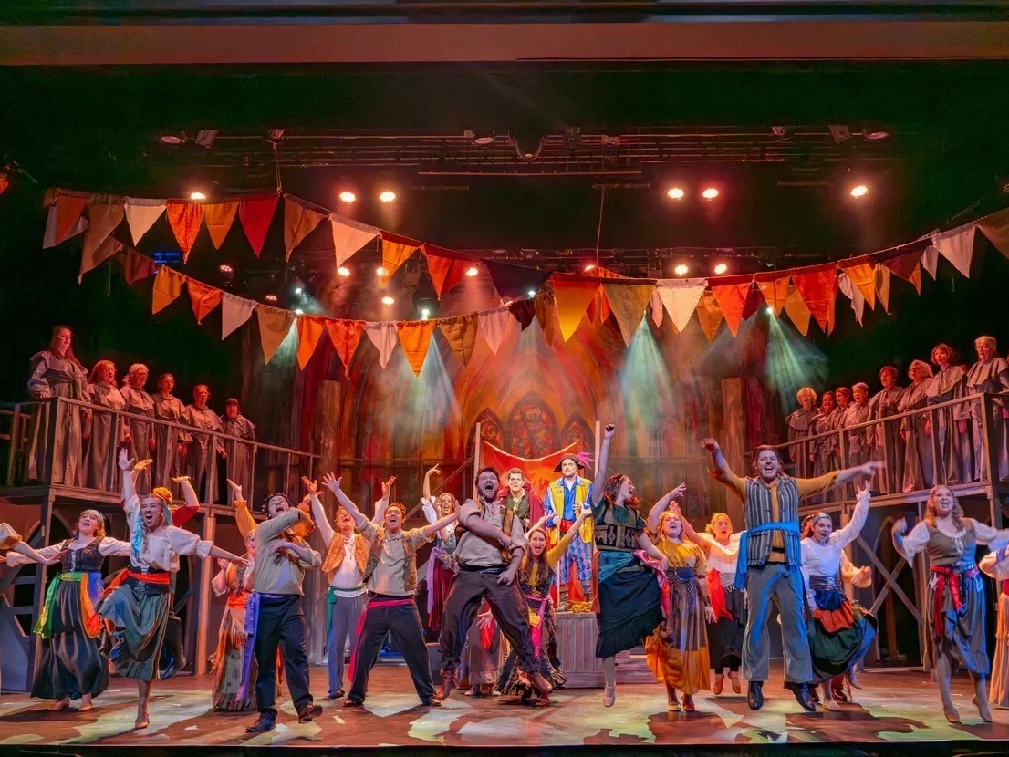 The image shows a lively stage performance with a large group of people dressed in colorful costumes. The stage is decorated with orange, yellow, and red bunting and features a wooden set with two levels. The performers are singing and dancing, with some raising their arms and others in mid-air, under warm stage lighting.