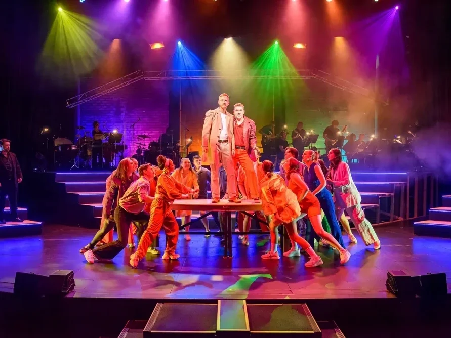 The stage scene from a musical theater performance with colorful lights illuminating the performers. Two male actors stand on a platform in the center, surrounded by cast members in vibrant costumes, with an orchestra playing in the background.