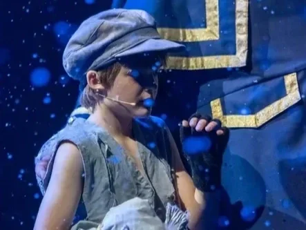 Child performing on stage with microphone, wearing a gray cap and costume, against a blue backdrop with stars.