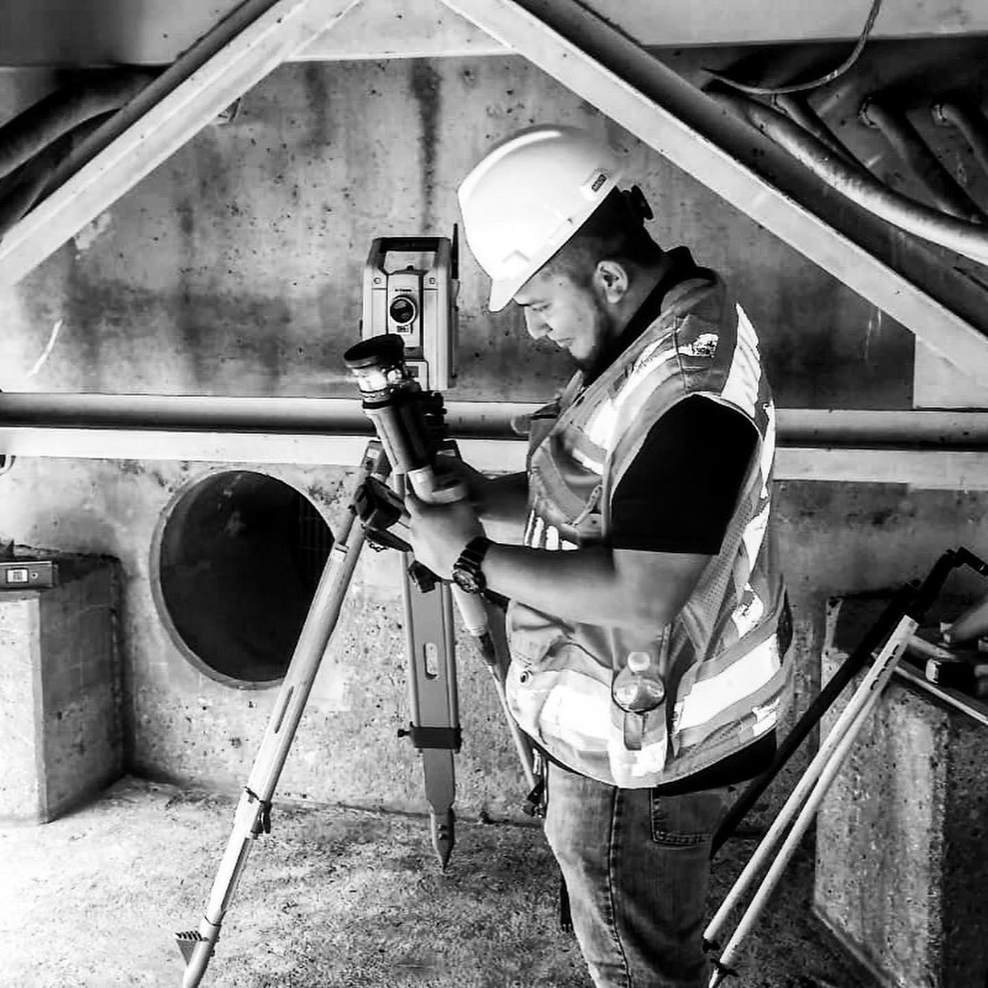 Construction worker wearing a safety vest and hard hat, looking at a surveying instrument on a tripod in a building under construction.