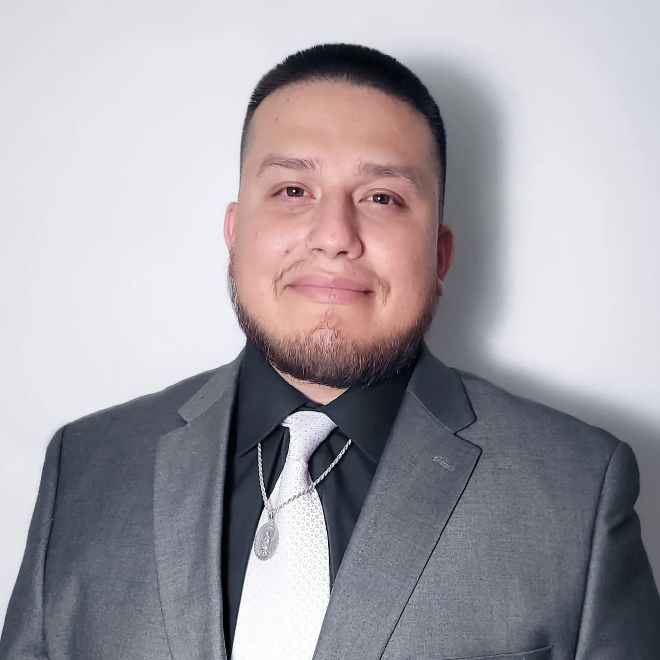 A young man with short black hair, a beard, and light skin wearing a dark gray suit, black shirt, and white tie, smiling against a plain white background.