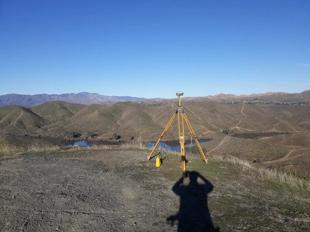 A surveying instrument set up on a tripod on a hilltop overlooking a valley with hills, a body of water, and dirt roads under a clear blue sky.