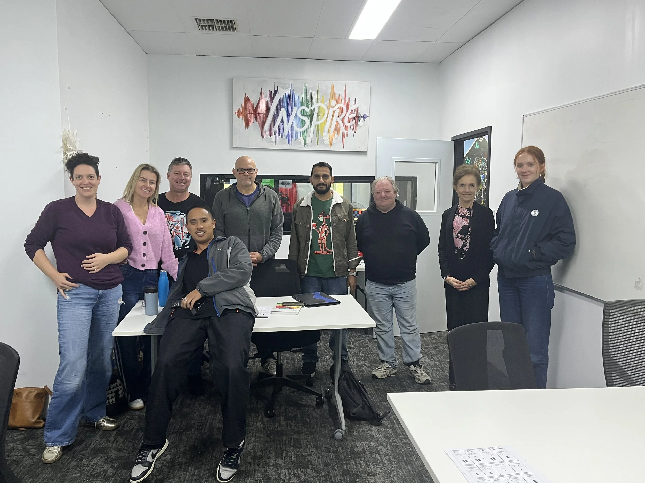 A group of nine people posing together for a photo in a meeting room, with a colorful sign that says 'INSPIRE' hanging on the wall behind them.
