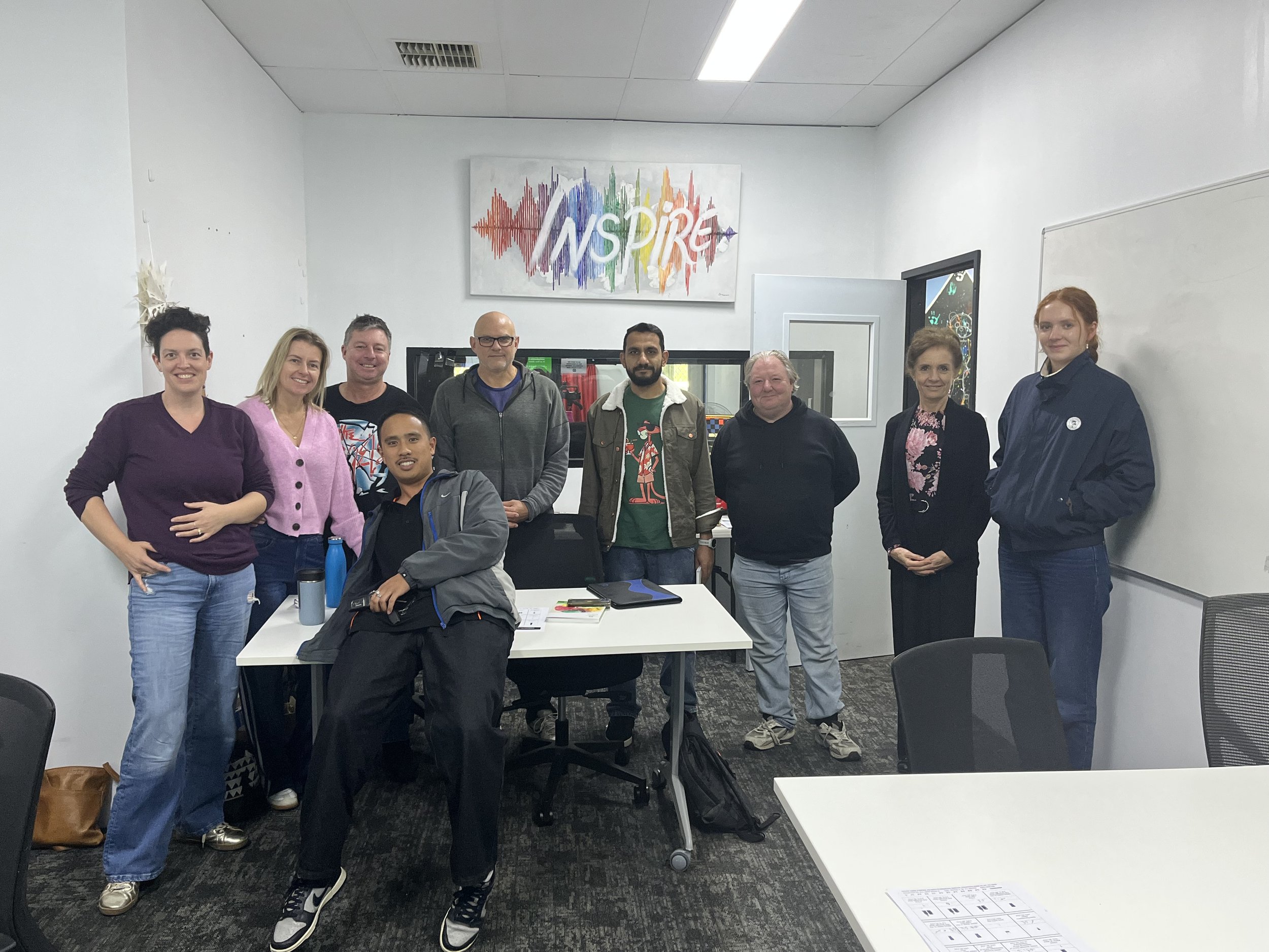 A group of nine people standing in a classroom or conference room with white walls and a whiteboard, posing for a photo. There is a colorful sign that says 'INSPIRE' on the wall behind them.