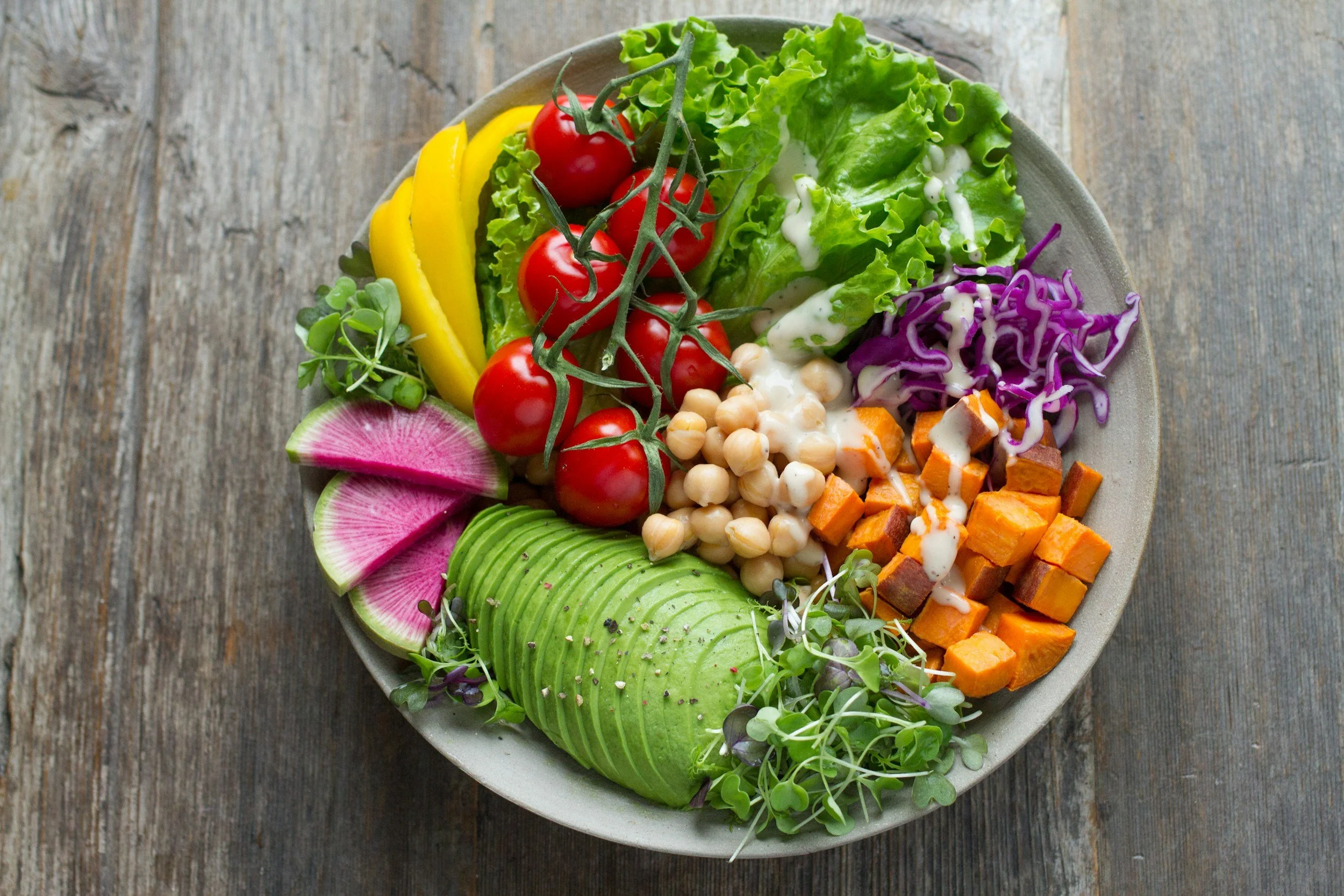 A bowl of mixed vegetables and salad, including cherry tomatoes, sliced radishes, avocado slices, shredded purple cabbage, diced sweet potatoes, chickpeas, and leafy greens.