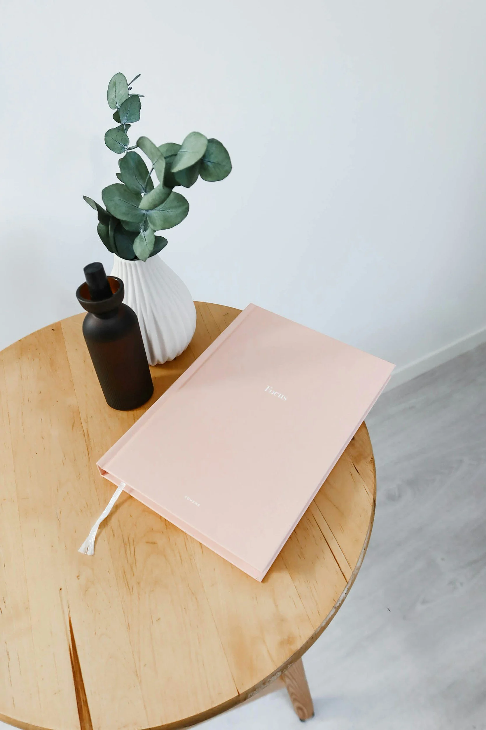 A wooden round table with a pink book, two vases (one black, one white) with green eucalyptus leaves, and a pink notebook branded 'focus' on a white background.