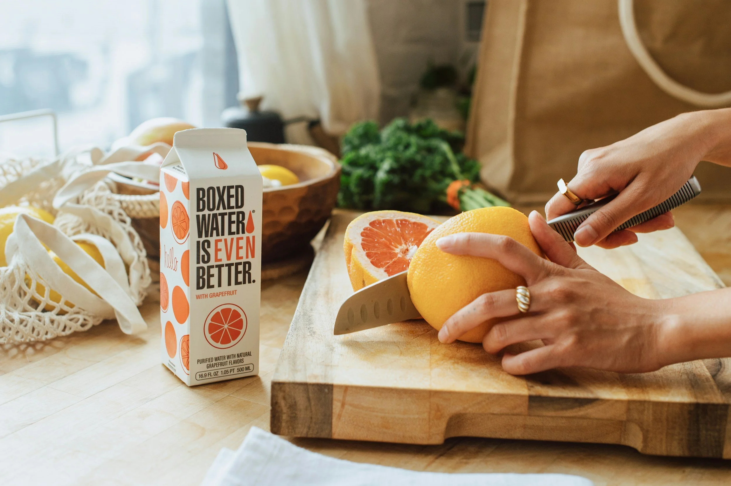 Person slicing a yellow grapefruit on a wooden cutting board in a kitchen, with a carton of boxed water with an orange slice graphic and the phrase "Boxed Water Is Even Better" nearby, and various fresh produce and a paper bag in the background.