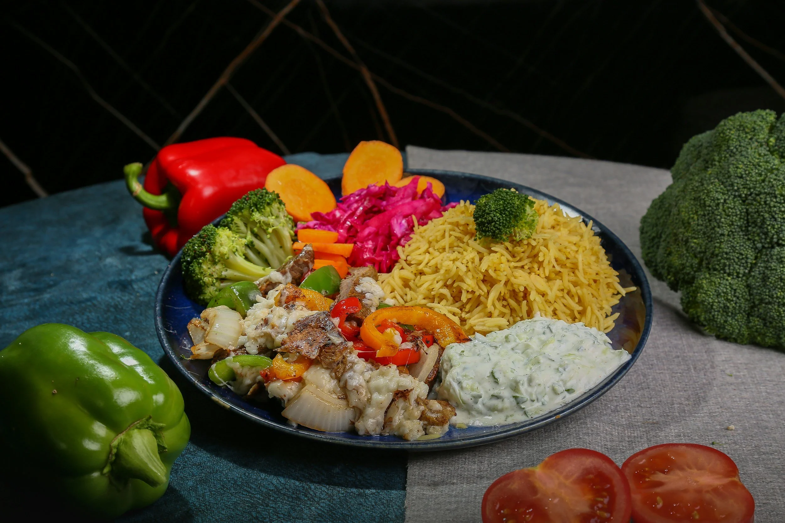 A bowl of cooked rice with broccoli, carrots, chopped meat, and a creamy sauce, surrounded by fresh vegetables including a red bell pepper, green bell pepper, broccoli, and sliced tomatoes on a gray table.