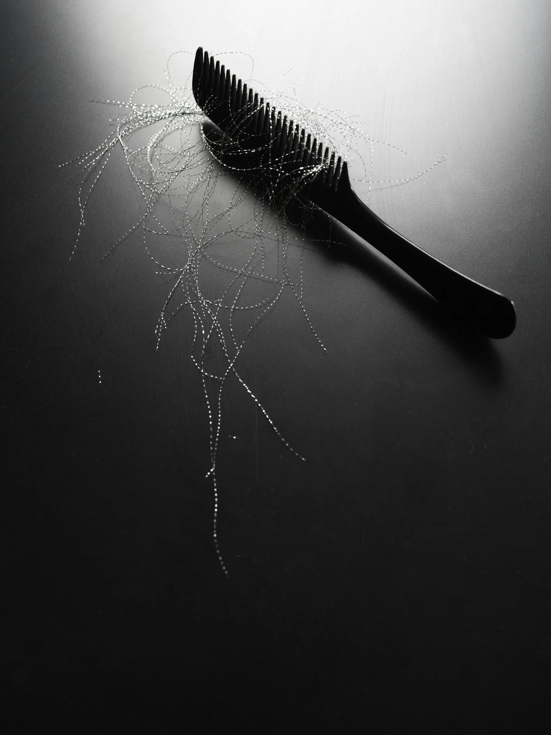 A black hair comb with many strands of silver thread tangled on and around it, placed on a dark surface.