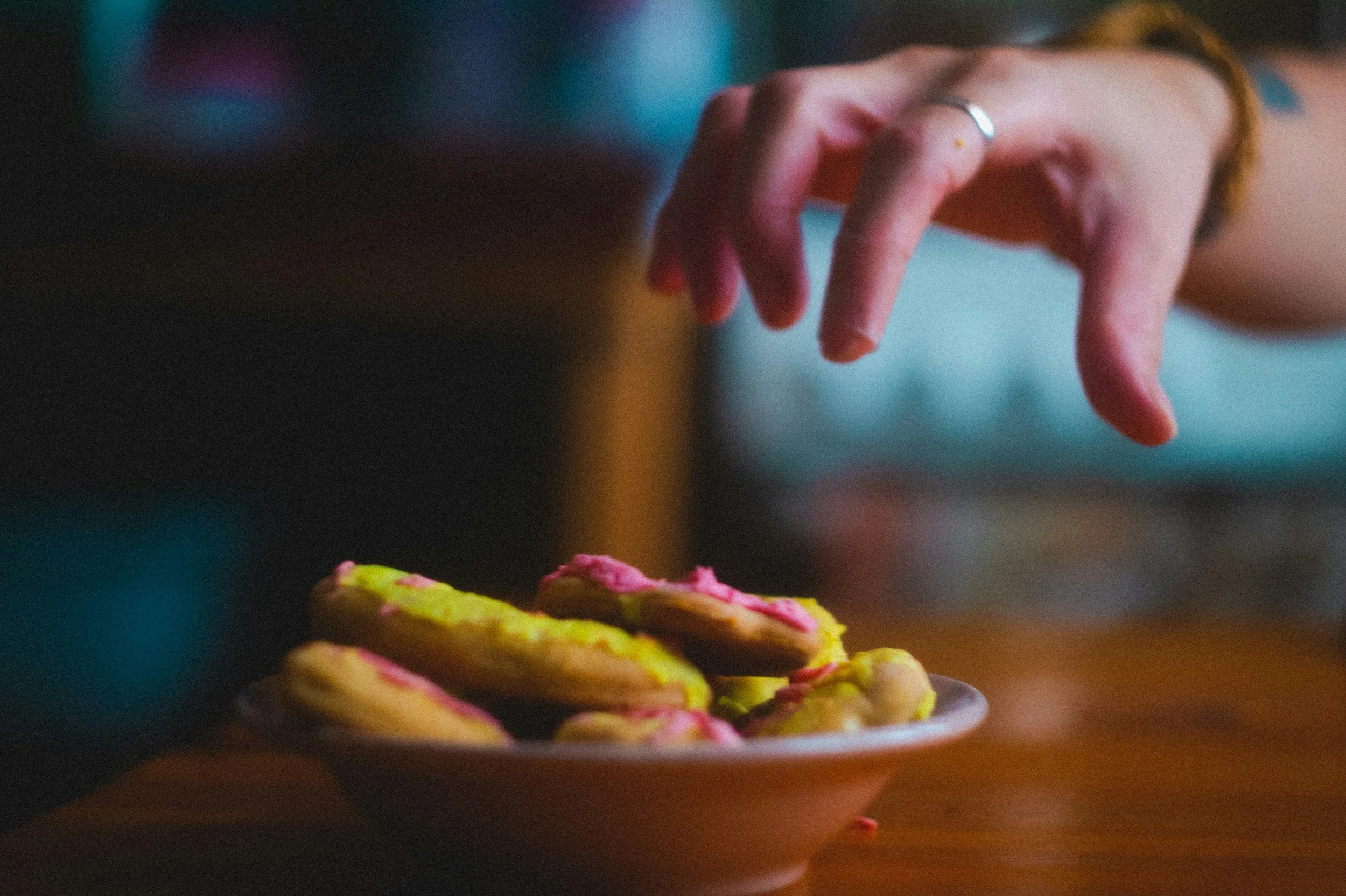 Close-up of a hand with a silver ring reaching to grab a cookie with pink icing and yellow edges on a plate. The background is blurry.
