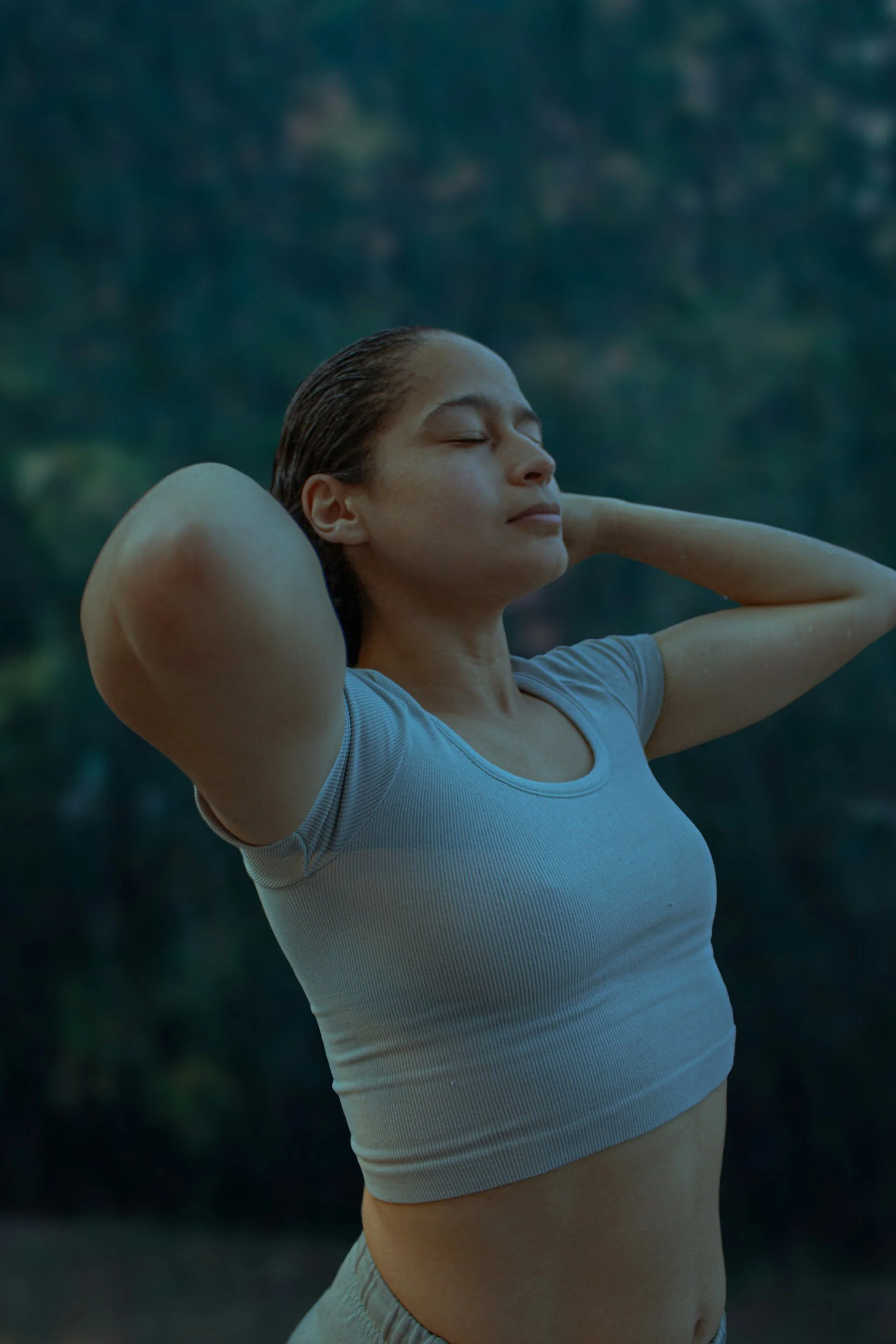 A woman with wet hair stretching outdoors against a background of trees and sky, wearing a light gray T-shirt and gray sweatpants.