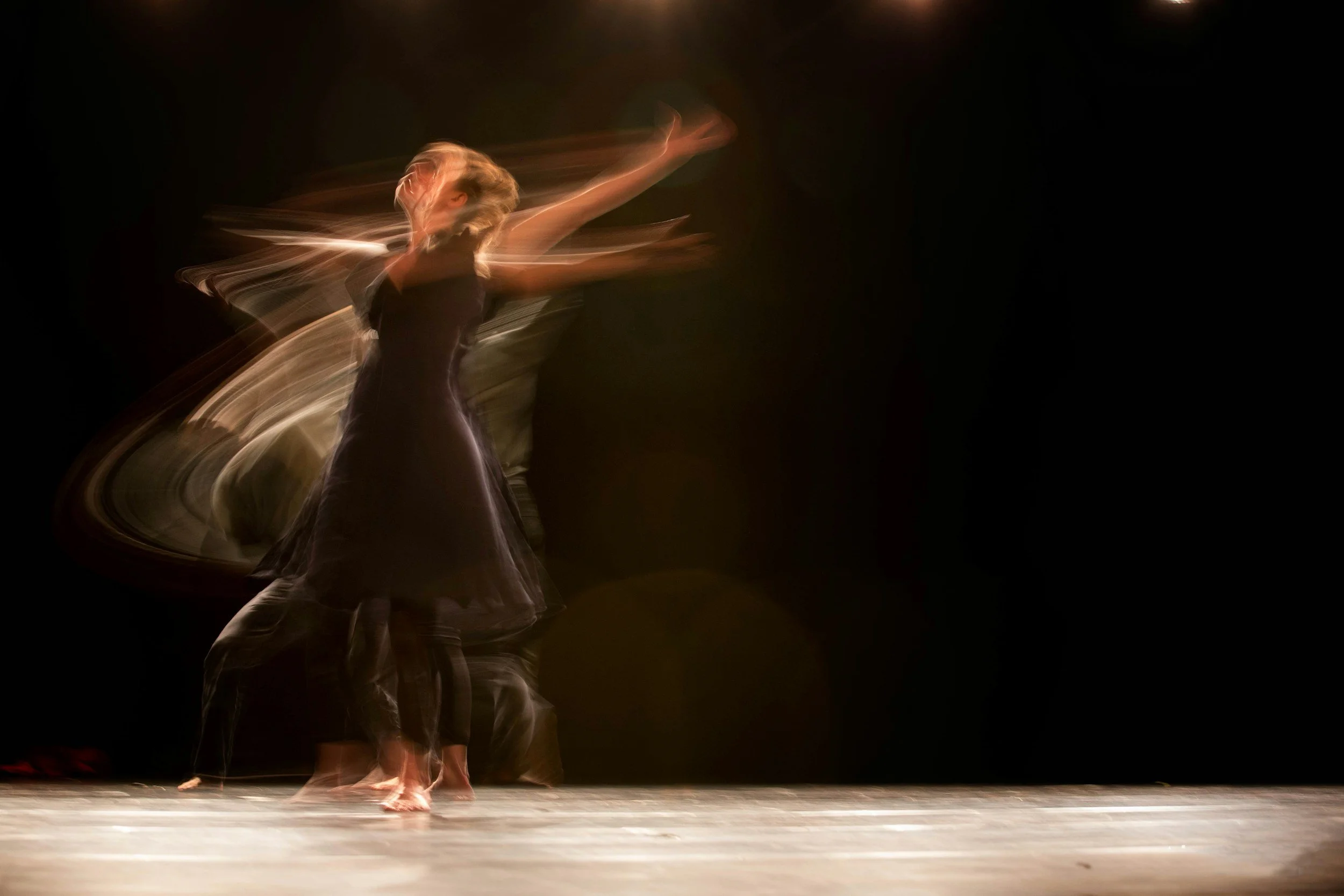 A dancer performing on a stage with motion blur effects, wearing a dark dress, with a dark background and wooden floor.