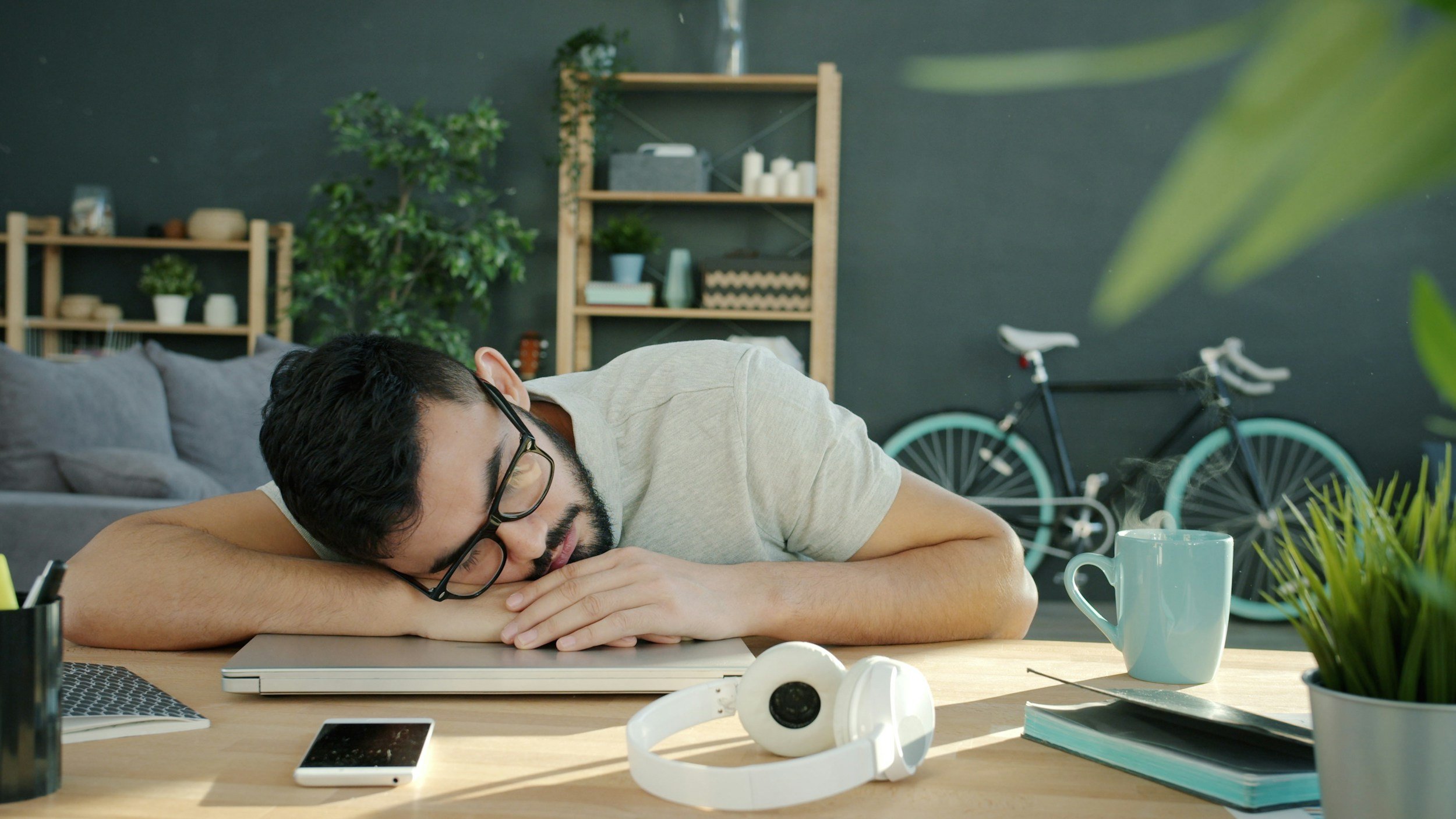 A man with glasses and a beard sleeping at a cluttered desk in a room with dark walls and wooden shelves, with a bicycle in the background.