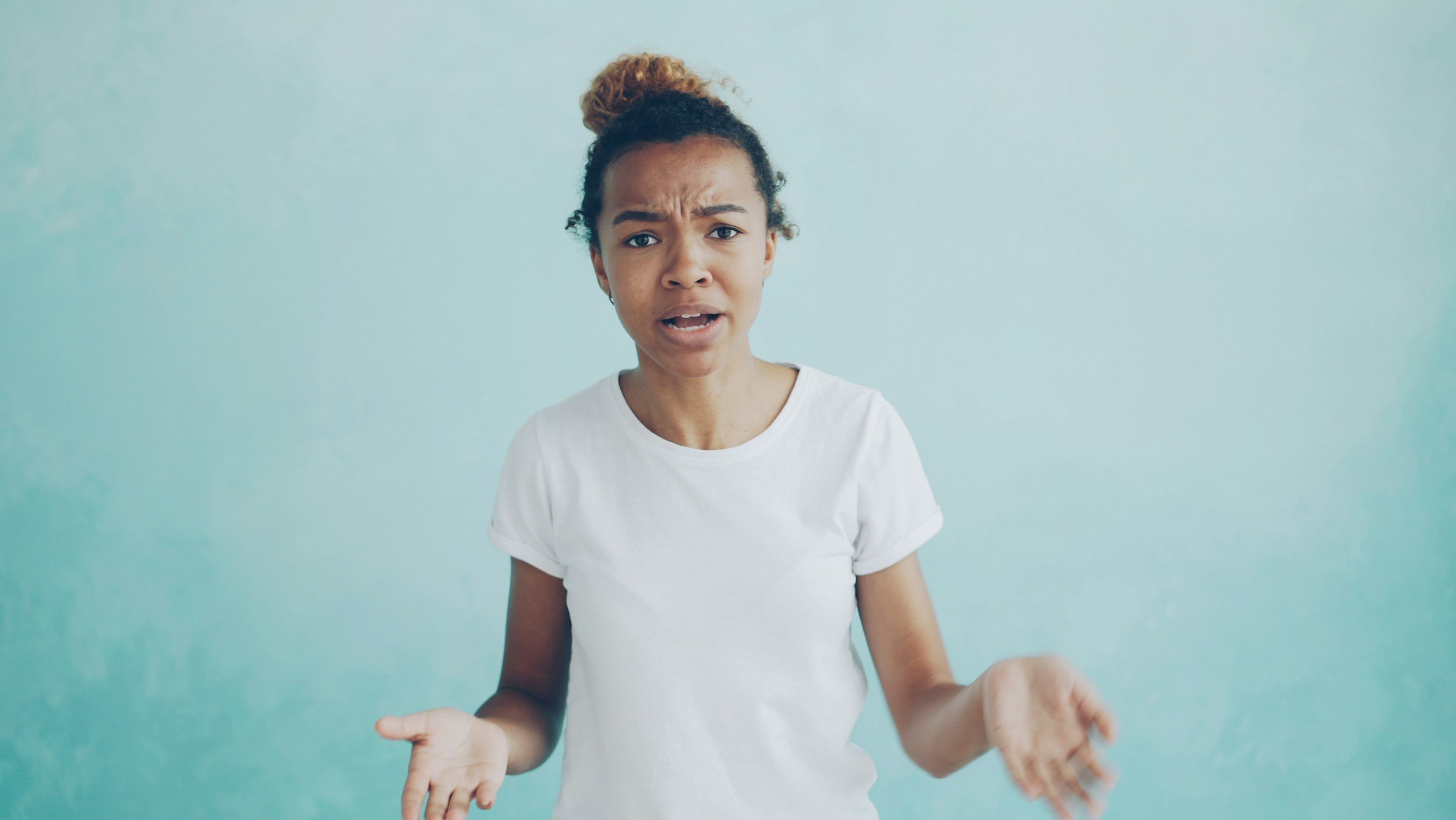 A young woman with curly hair and a surprised expression, wearing a white t-shirt, standing against a light blue wall.