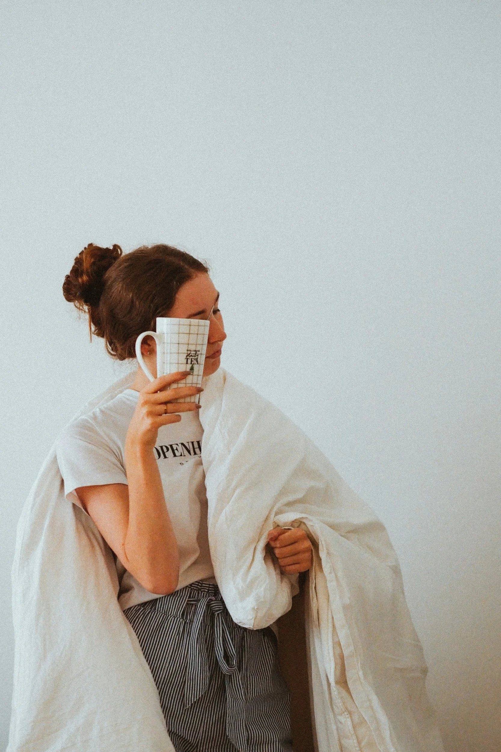 A woman with brown hair in a bun holding a coffee mug with a grid pattern, standing next to a person wrapped in a beige blanket, against a plain white wall.