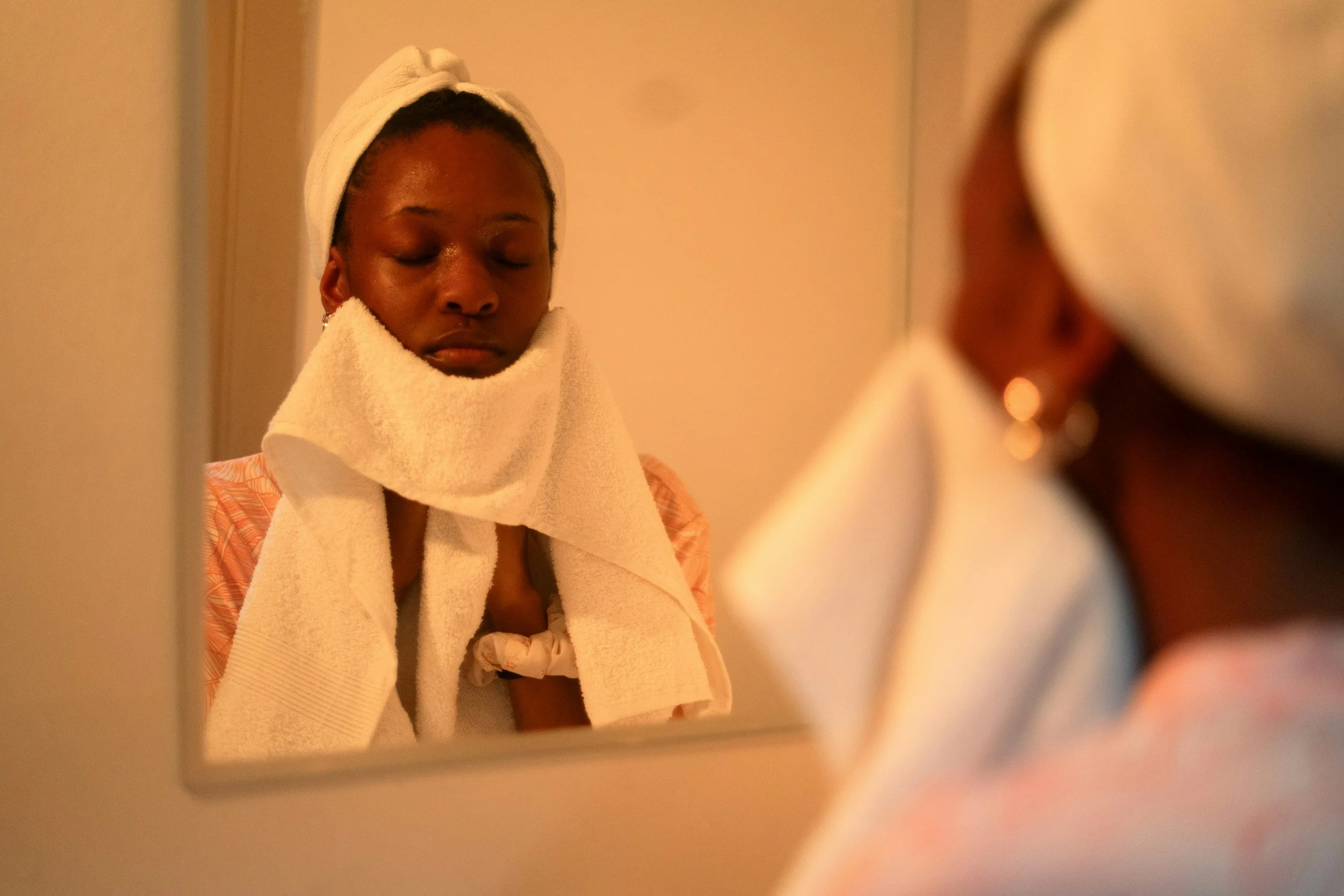 A young woman with dark skin and closed eyes, wearing a towel wrapped around her head and a bathrobe, looking at her reflection in a mirror.