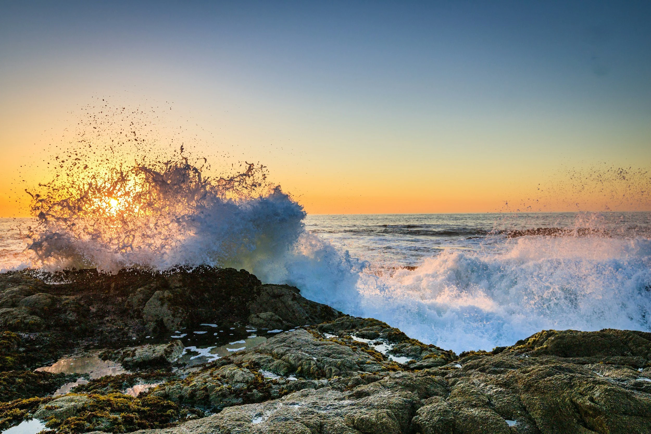 Sunset over the ocean with waves crashing against rocks and the sky transitioning from orange to blue.