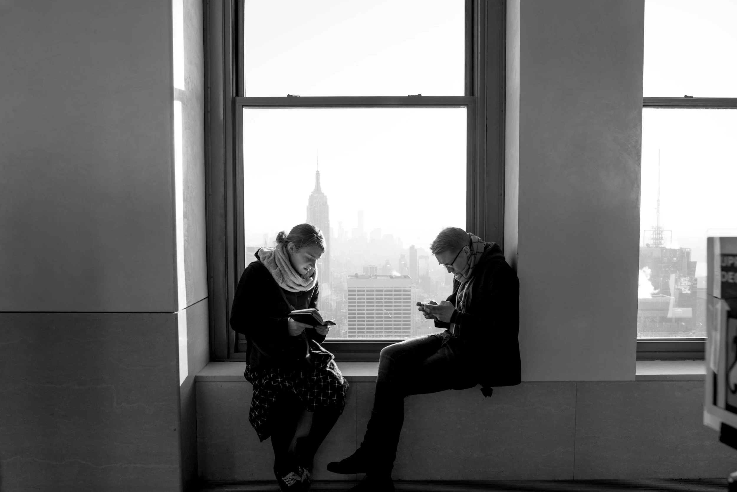 Two people sitting by a large window in a high-rise building, looking at their phones, with the New York City skyline, including the Empire State Building, visible in the background.