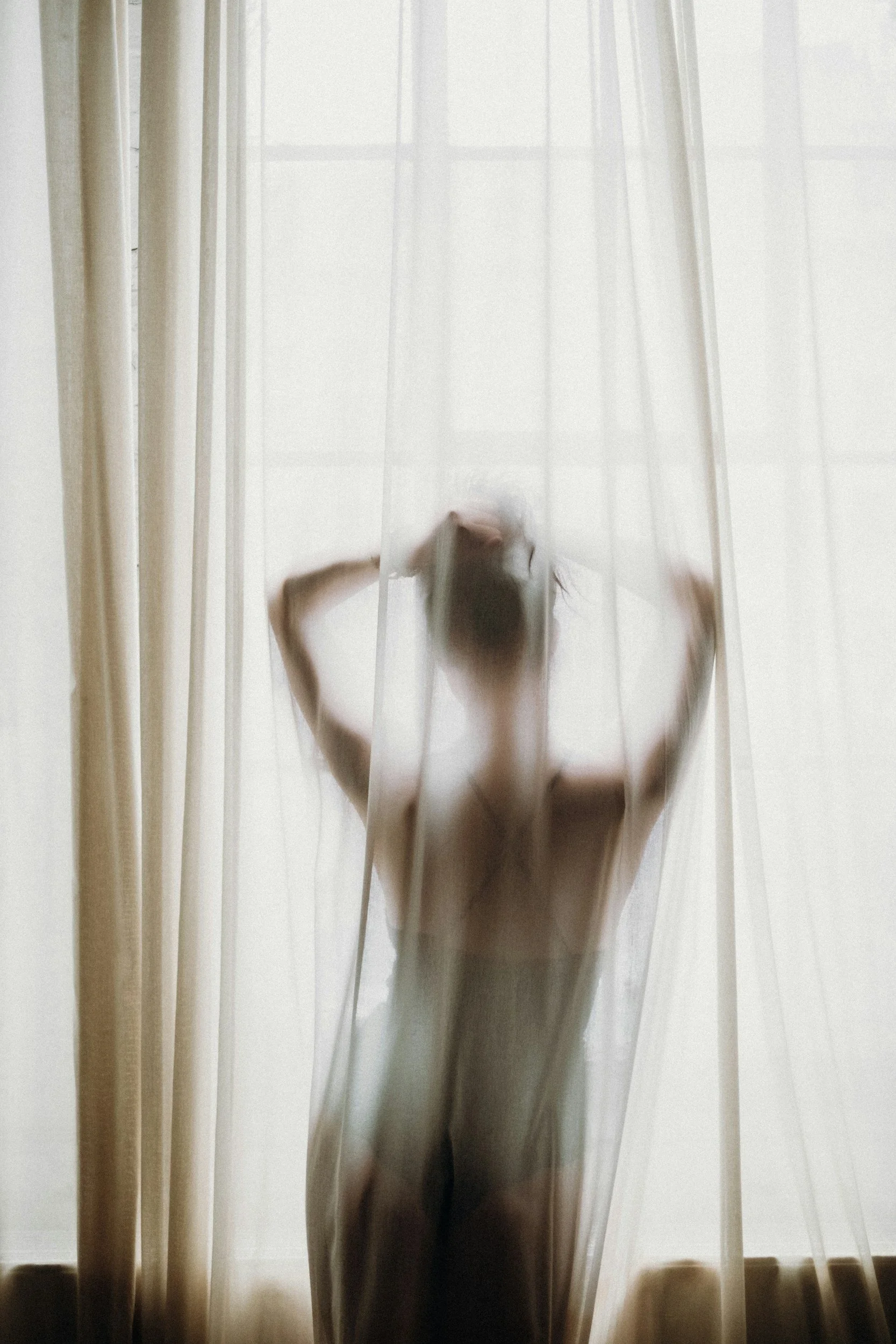 A person standing behind sheer curtains near a window, with their arms raised and head slightly bent, creating a blurred silhouette.
