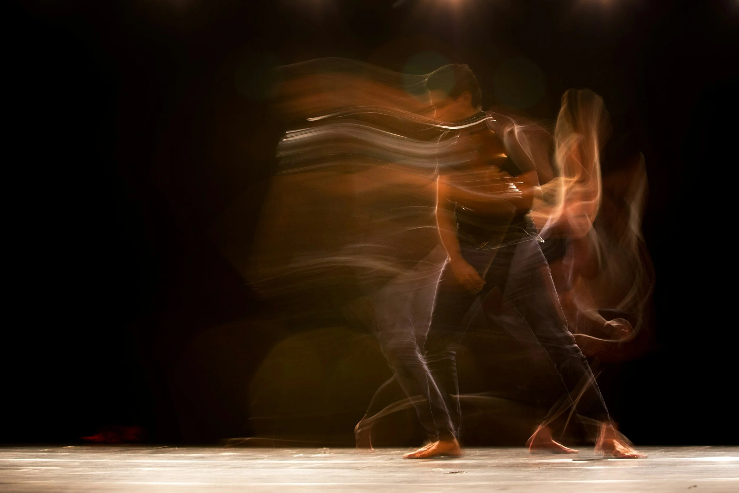 A person dancing on stage with a dark background and wooden floor, captured with motion blur to show movement and energy.