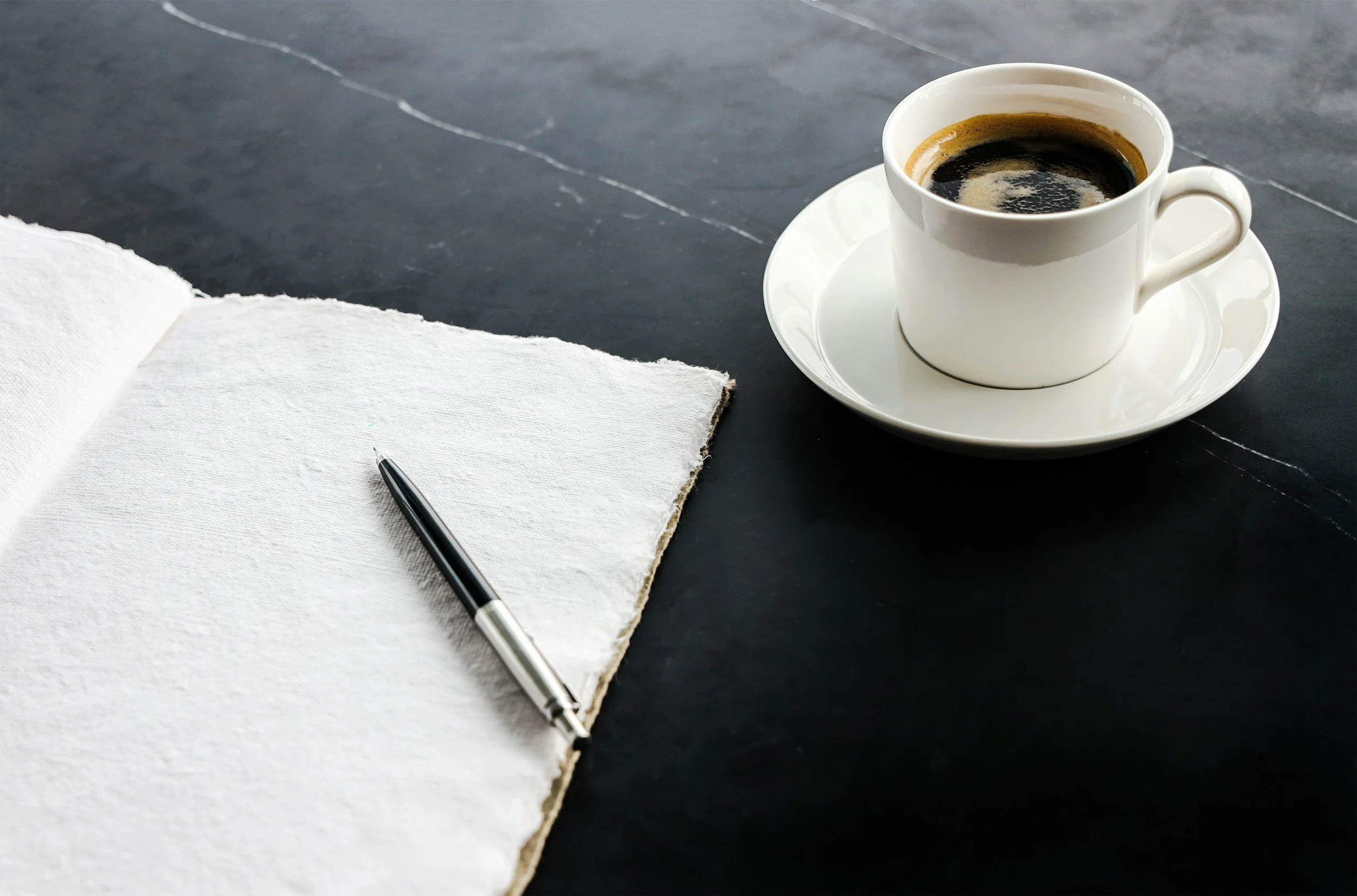 A white coffee cup filled with black coffee on a matching saucer, an open notebook with a black pen resting on it, all placed on a dark slate surface.