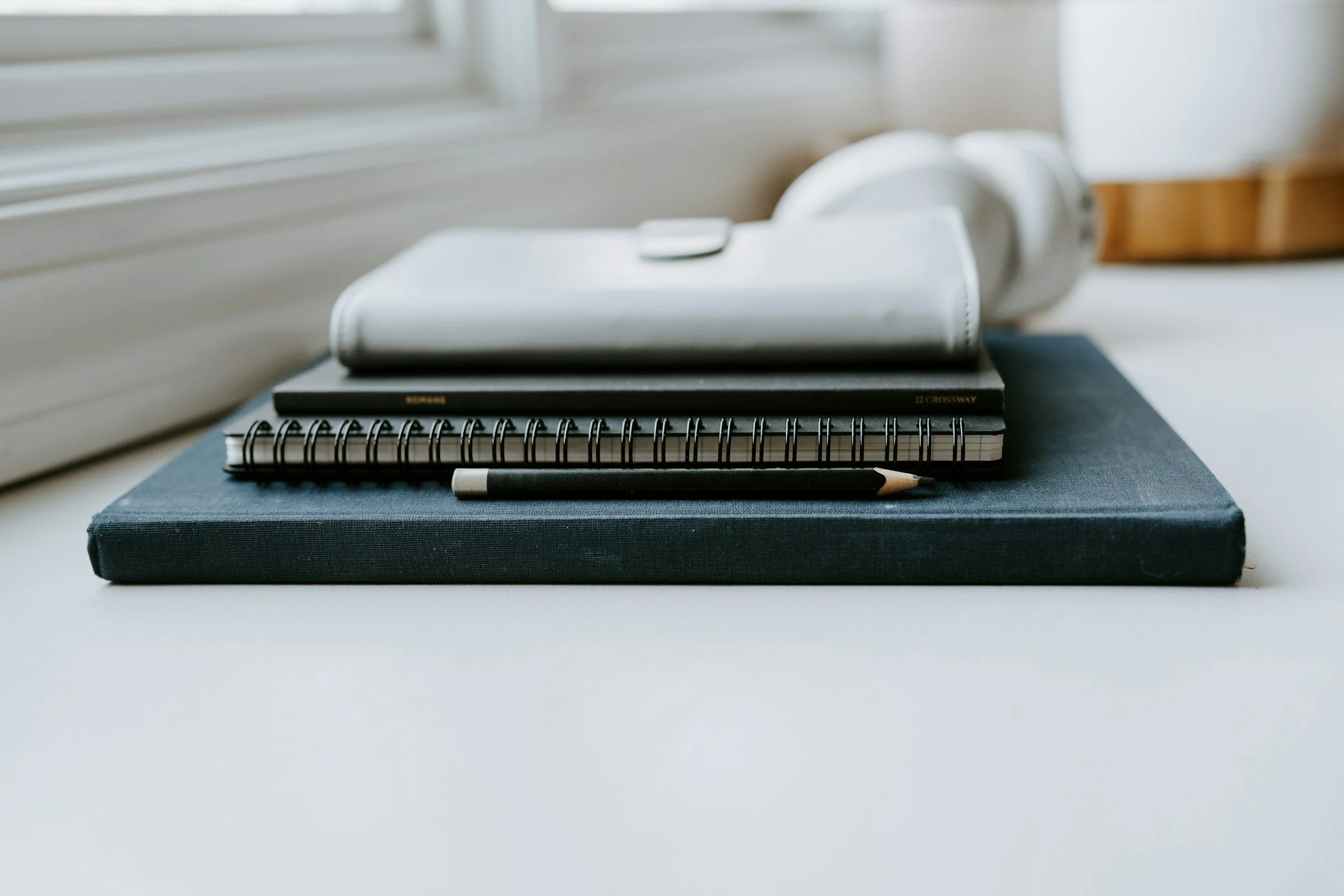 A white cordless vacuum cleaner resting on top of a closed black notebook with a spiral binding, a black notebook, and a black pen on a light-colored surface near a window with closed blinds.