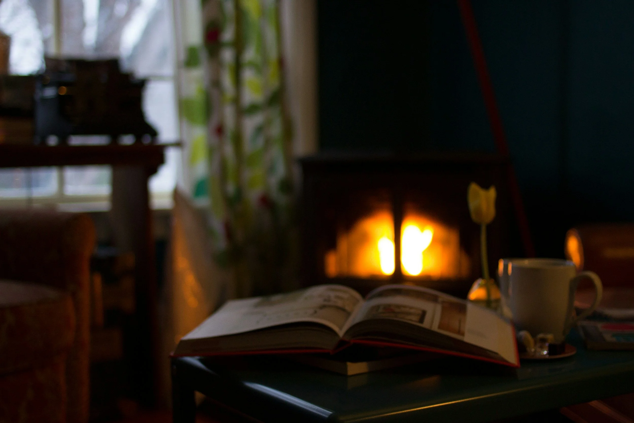 A cozy indoor scene with a table, a lamp, an open magazine, a coffee cup, a yellow flower, and a window with curtains. The background features a dark wall with a warm glow.