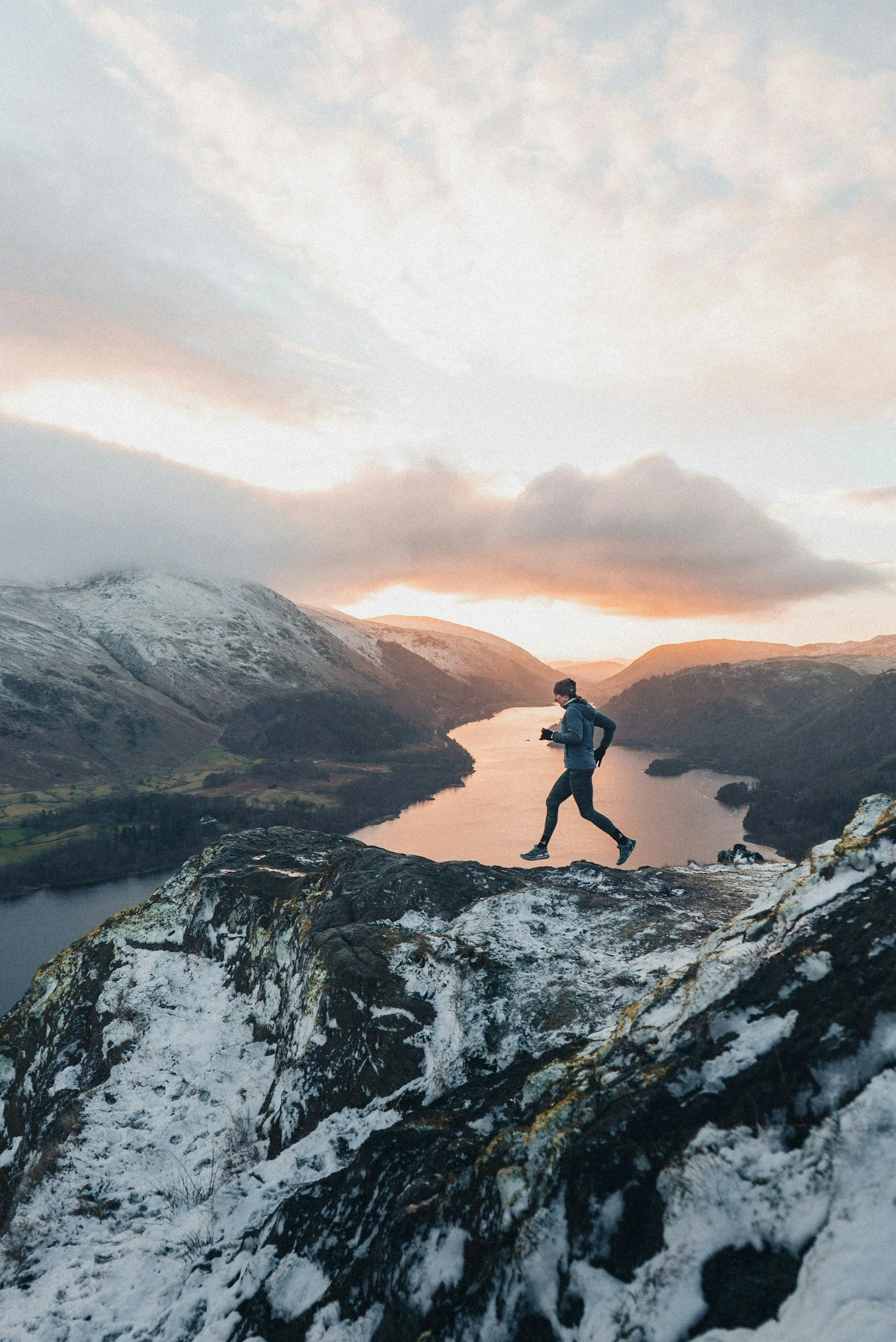 A person running on a rocky trail overlooking a landscape of mountains, a lake, and a cloudy sky at sunset.