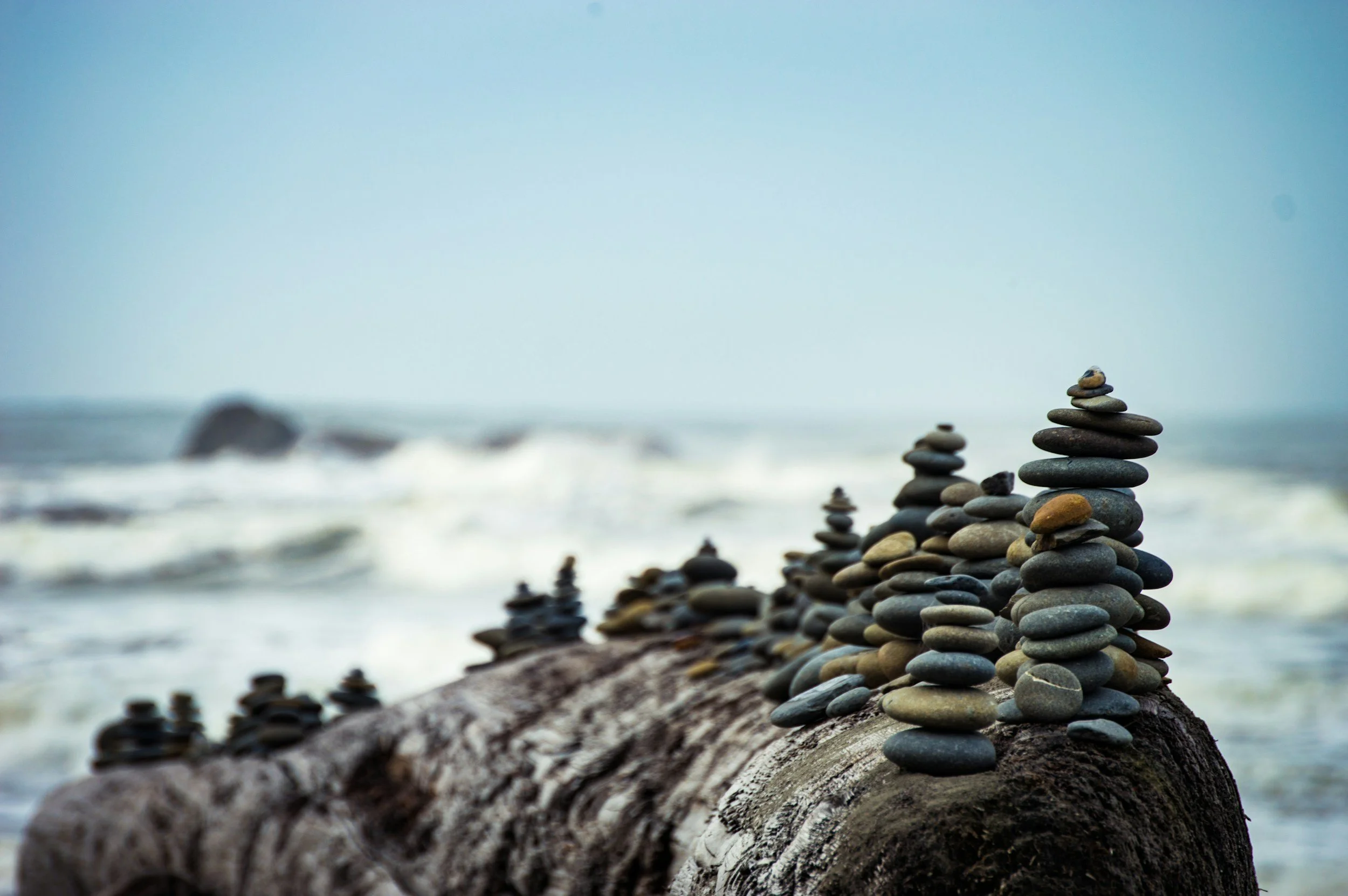 Stacks of small smooth stones arranged on driftwood on a beach, with the ocean and sky in the background.