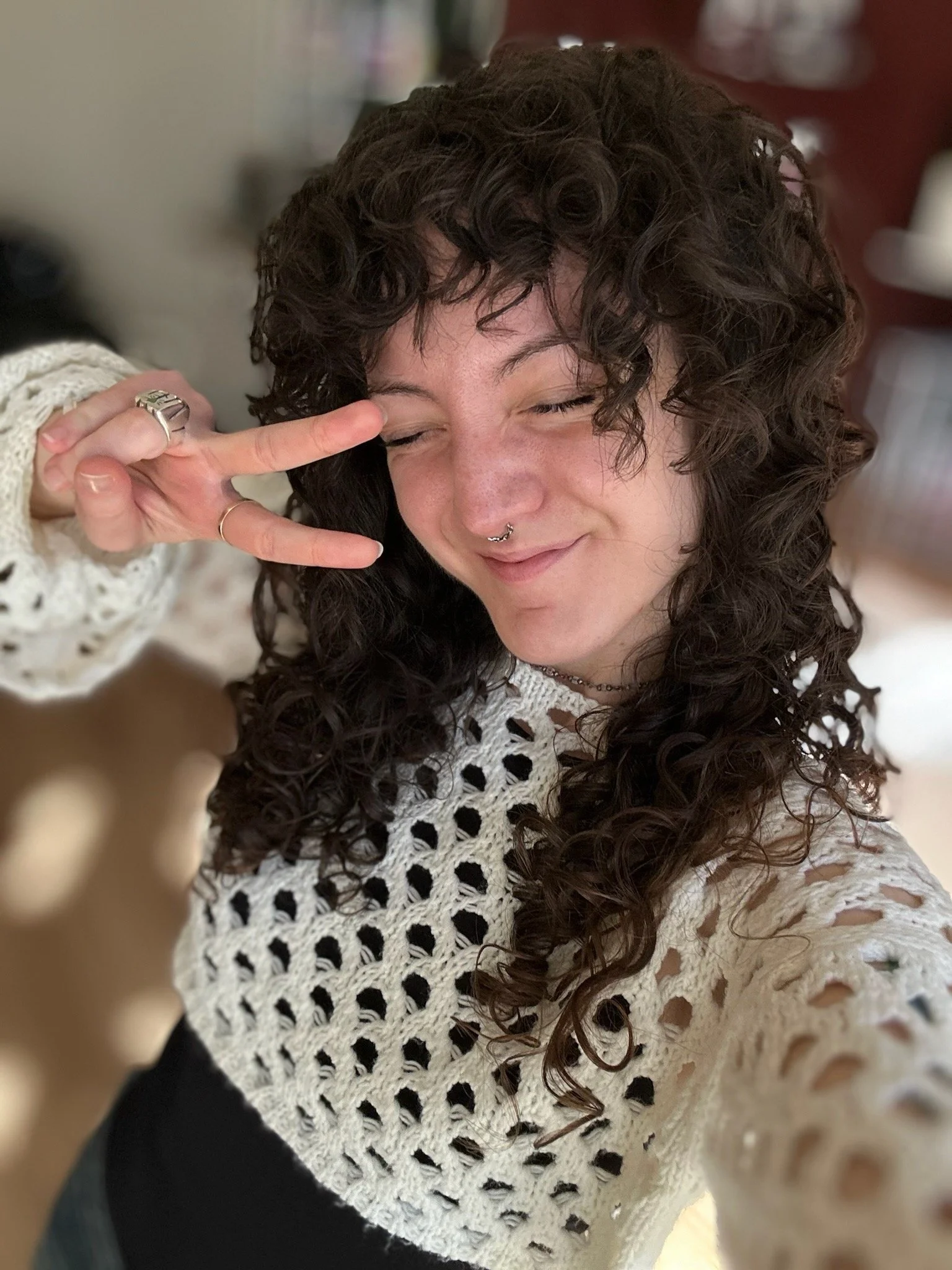 Close-up of a woman with curly brown hair smiling and making a peace sign with her hand near her face. She is wearing a white crocheted top and rings on her fingers.