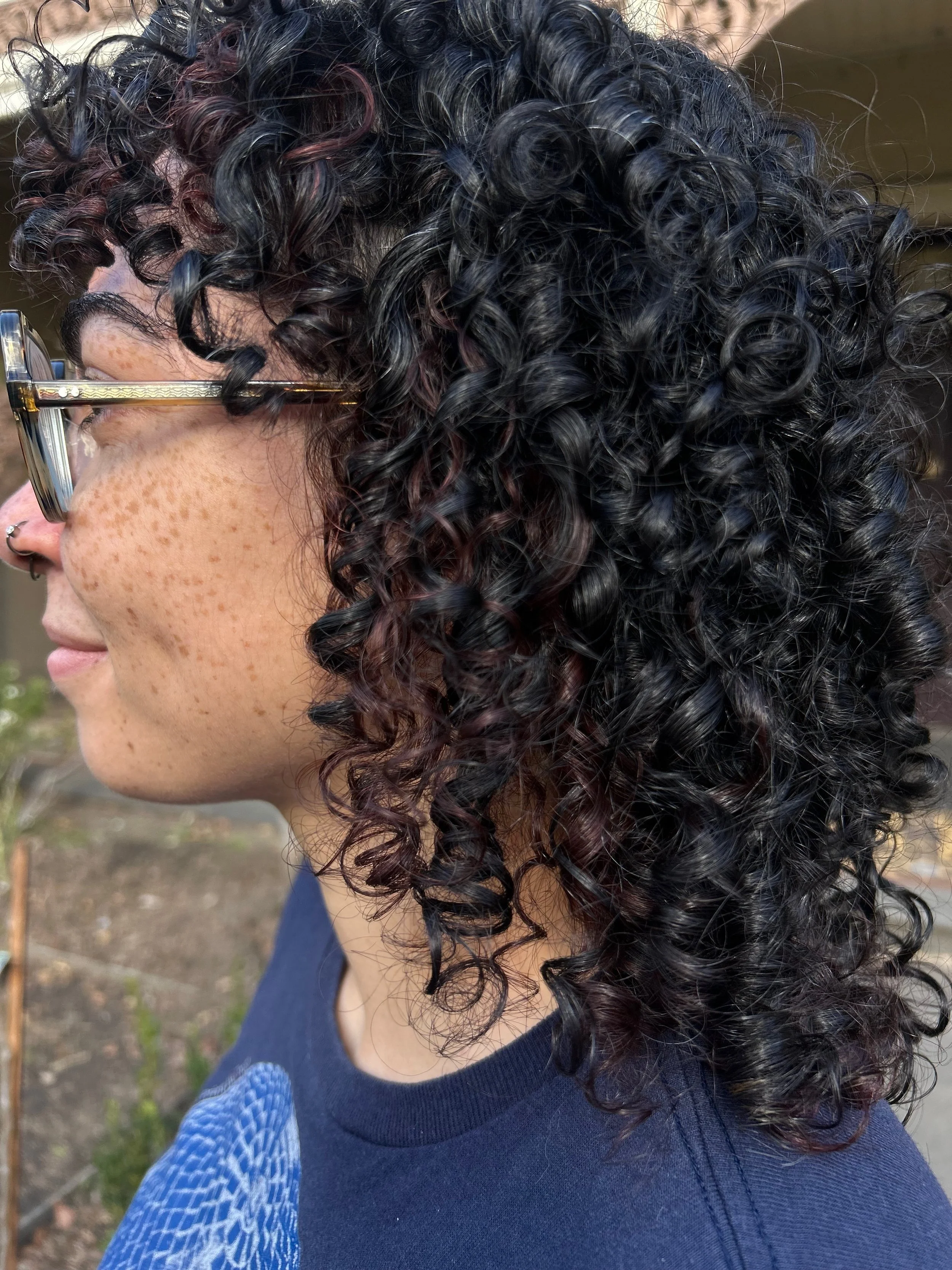 A person with curly dark hair and glasses, wearing a dark blue shirt, appears to be outdoors during daytime.
