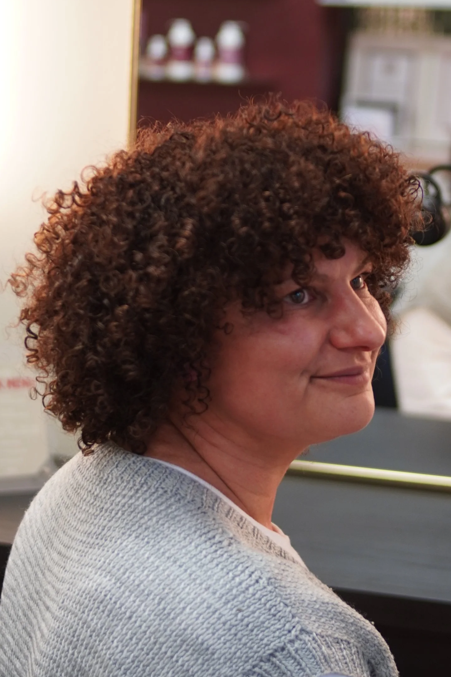 A woman with curly brown hair and fair skin, wearing a light gray sweater, sitting in an indoor setting.