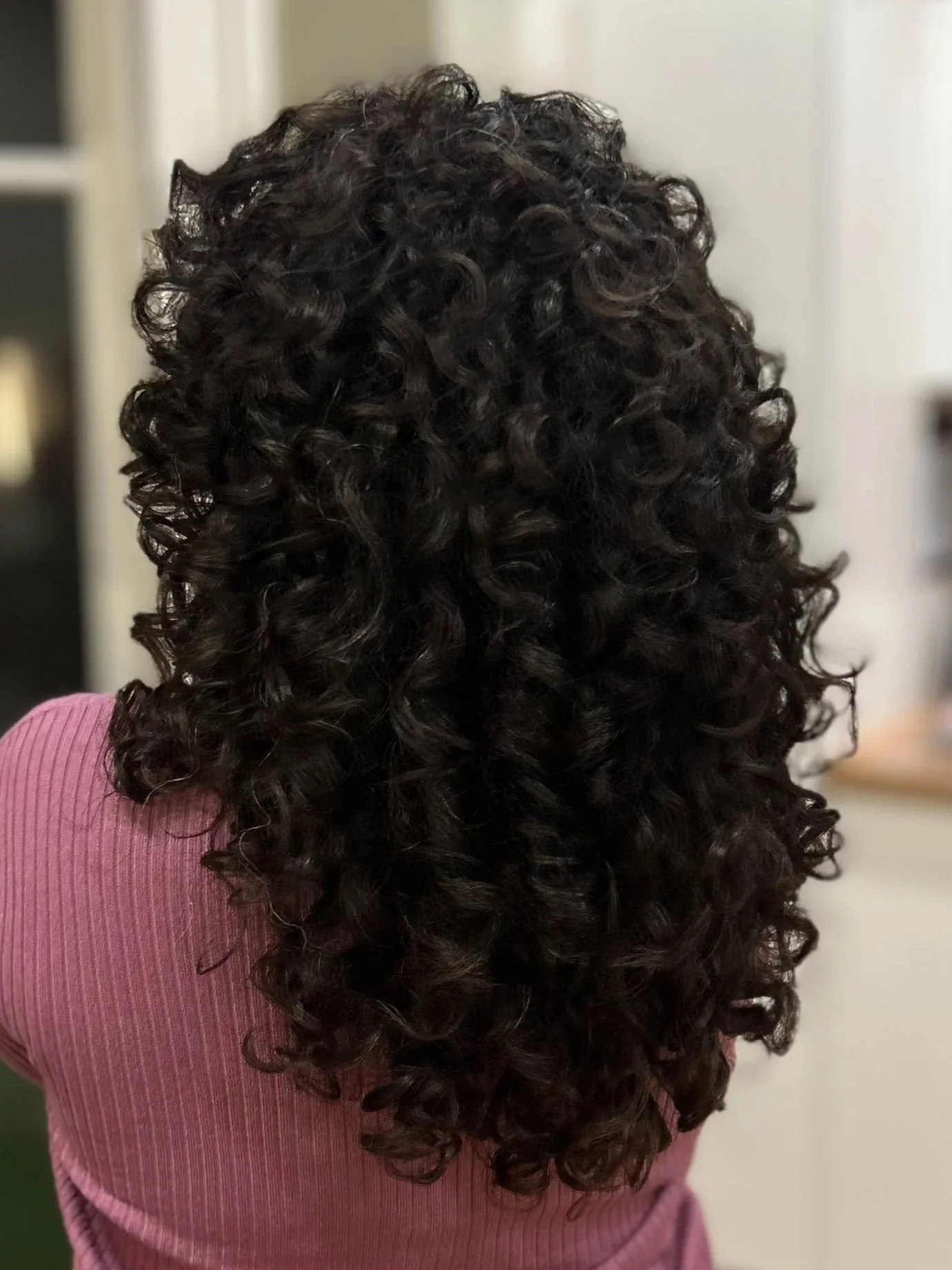 Back view of a person with long, dark, curly hair wearing a pink top.