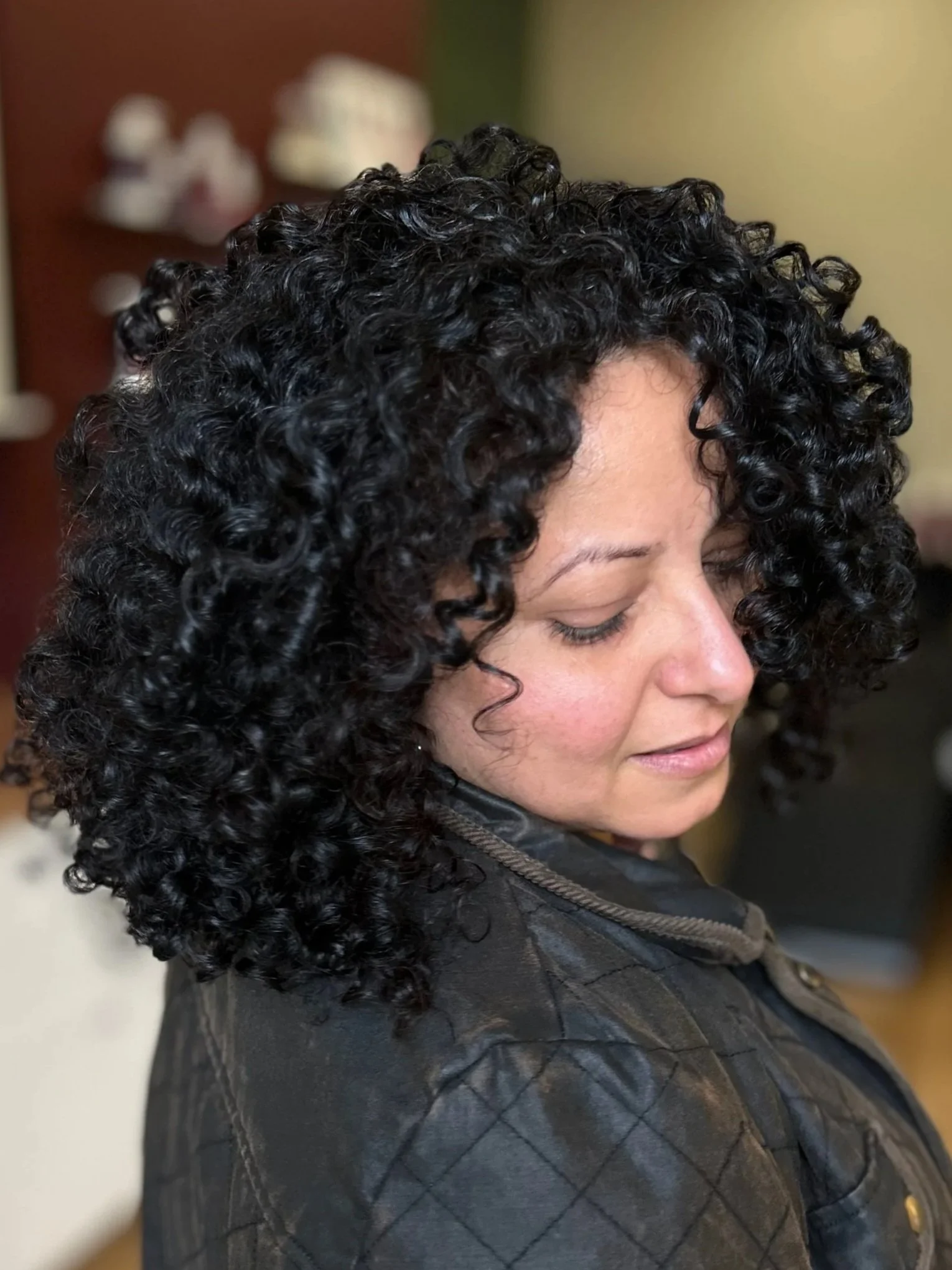 A woman with dark, curly hair, wearing a black quilted jacket, with her eyes closed, captured in profile.