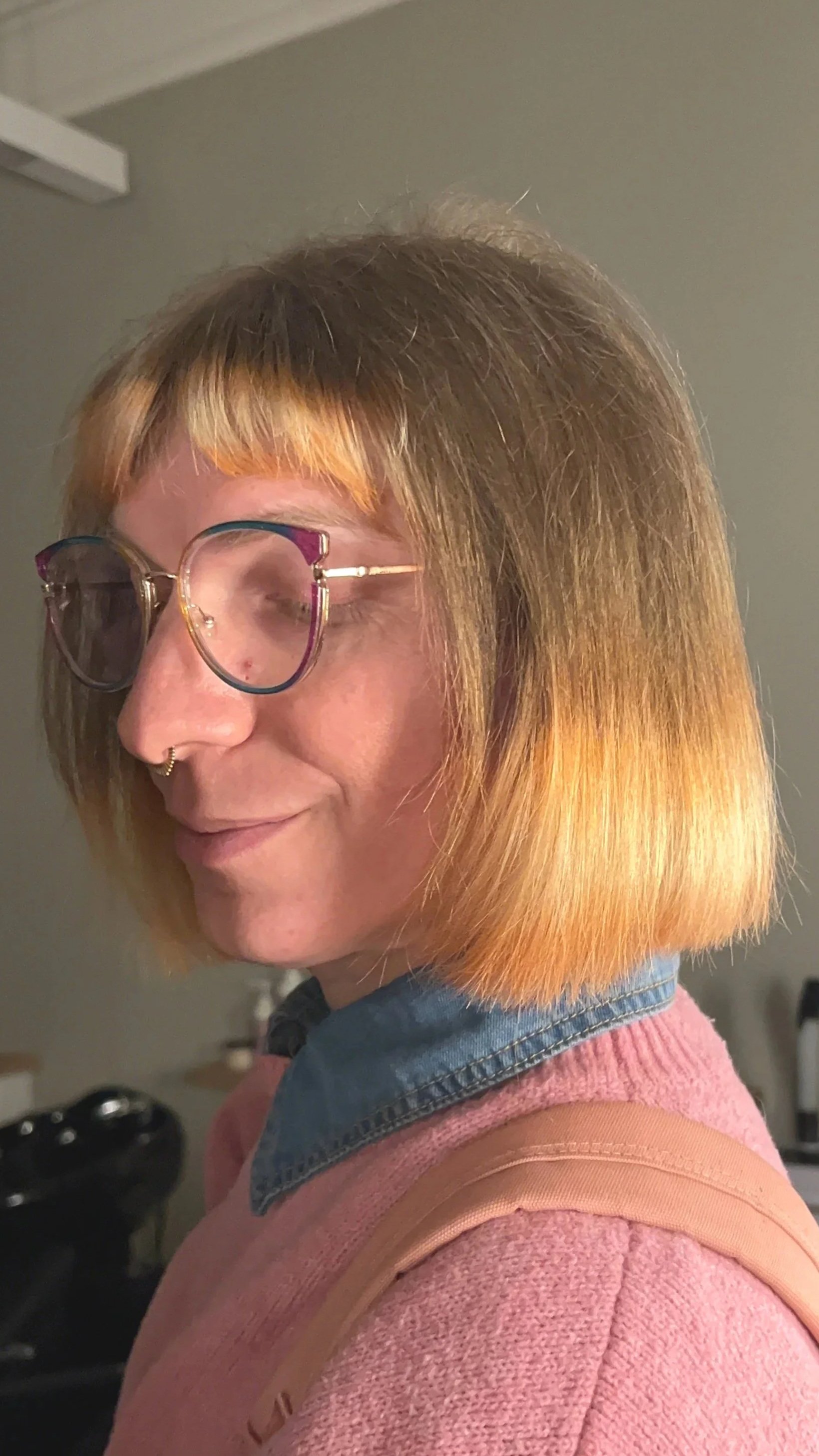 A woman with short, two-tone hair (brown with orange highlights) wearing glasses, a nose ring, a pink sweater, and a denim jacket.