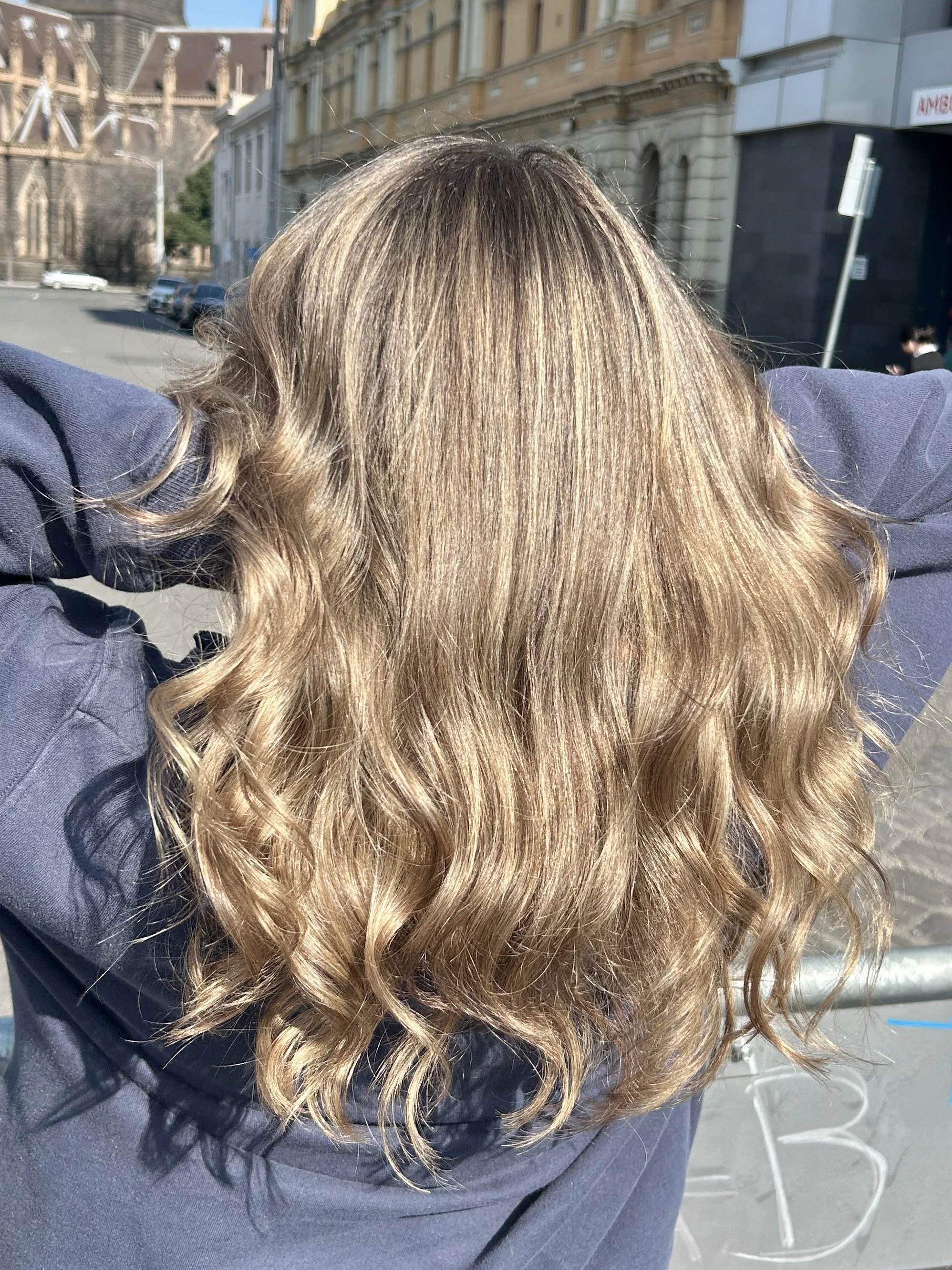 Person with long, curly blonde hair wearing a dark sweatshirt outdoors on a sunny day.
