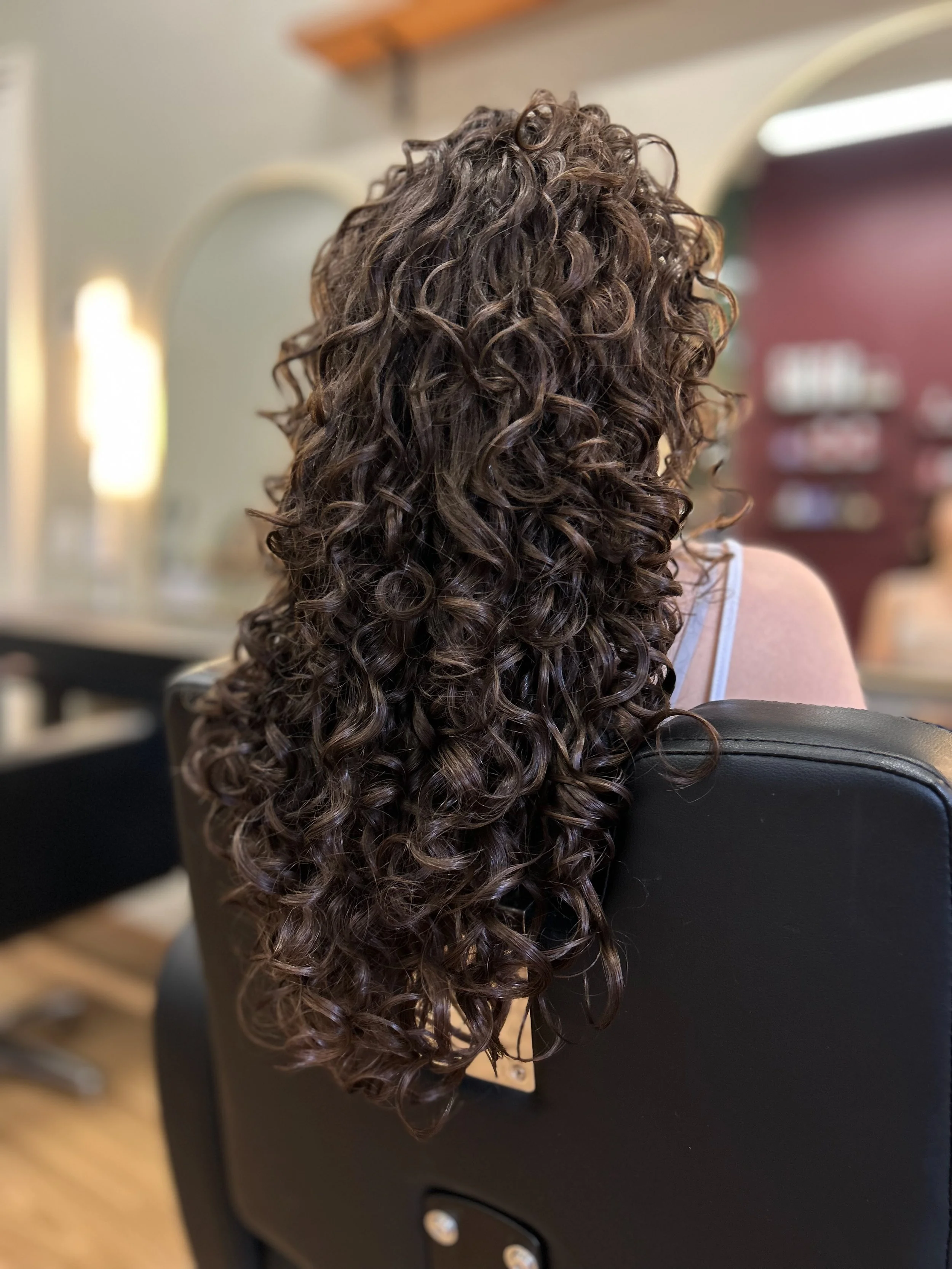 Back view of a woman with long, curly brown hair sitting in a black salon chair, with the focus on her hair and blurred salon background.