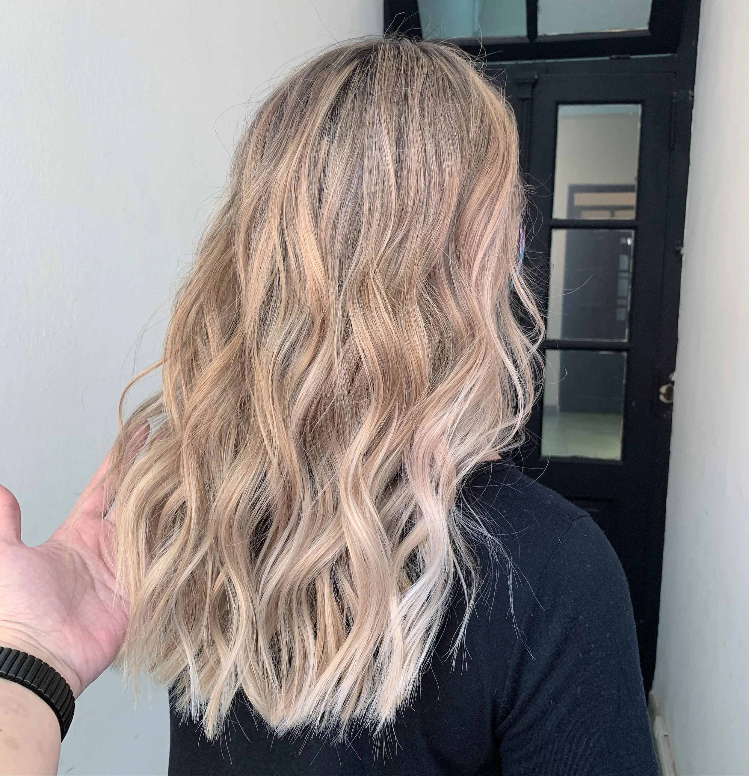 A person with long, wavy blonde hair, wearing a pink garment on their shoulder, a black wristwatch, and a black shirt. Another person's hand with a black bracelet is touching their hair. The background includes a white wall and a black door with glass panels.
