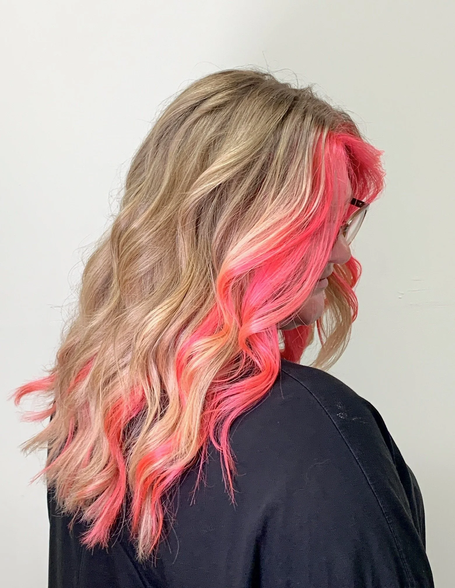 Side view of a woman with long blonde hair with pink highlights and curls, dressed in a black top, against a plain white background.