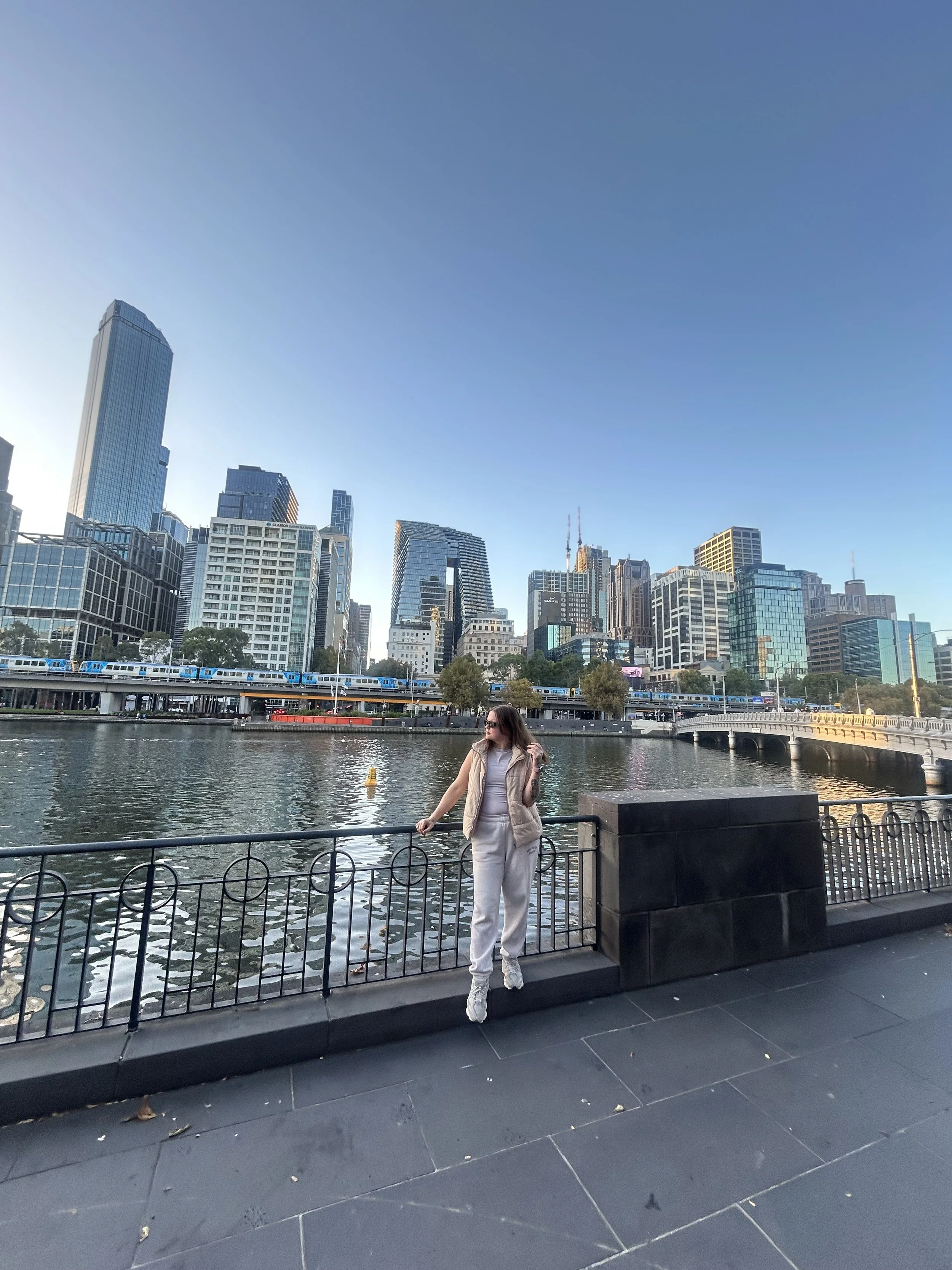 A young woman standing at a railing by a river in a city. The background features a skyline with tall skyscrapers, including a prominent glass building, under a clear blue sky, with a train passing over a bridge.