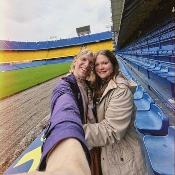 Two women taking a selfie at a baseball stadium with vacant blue seats and a dirt field.