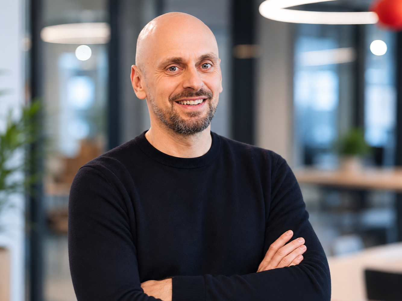 A smiling man with a beard and shaved head, wearing a black sweater, standing with arms crossed in a modern office environment with large windows and hanging lights.