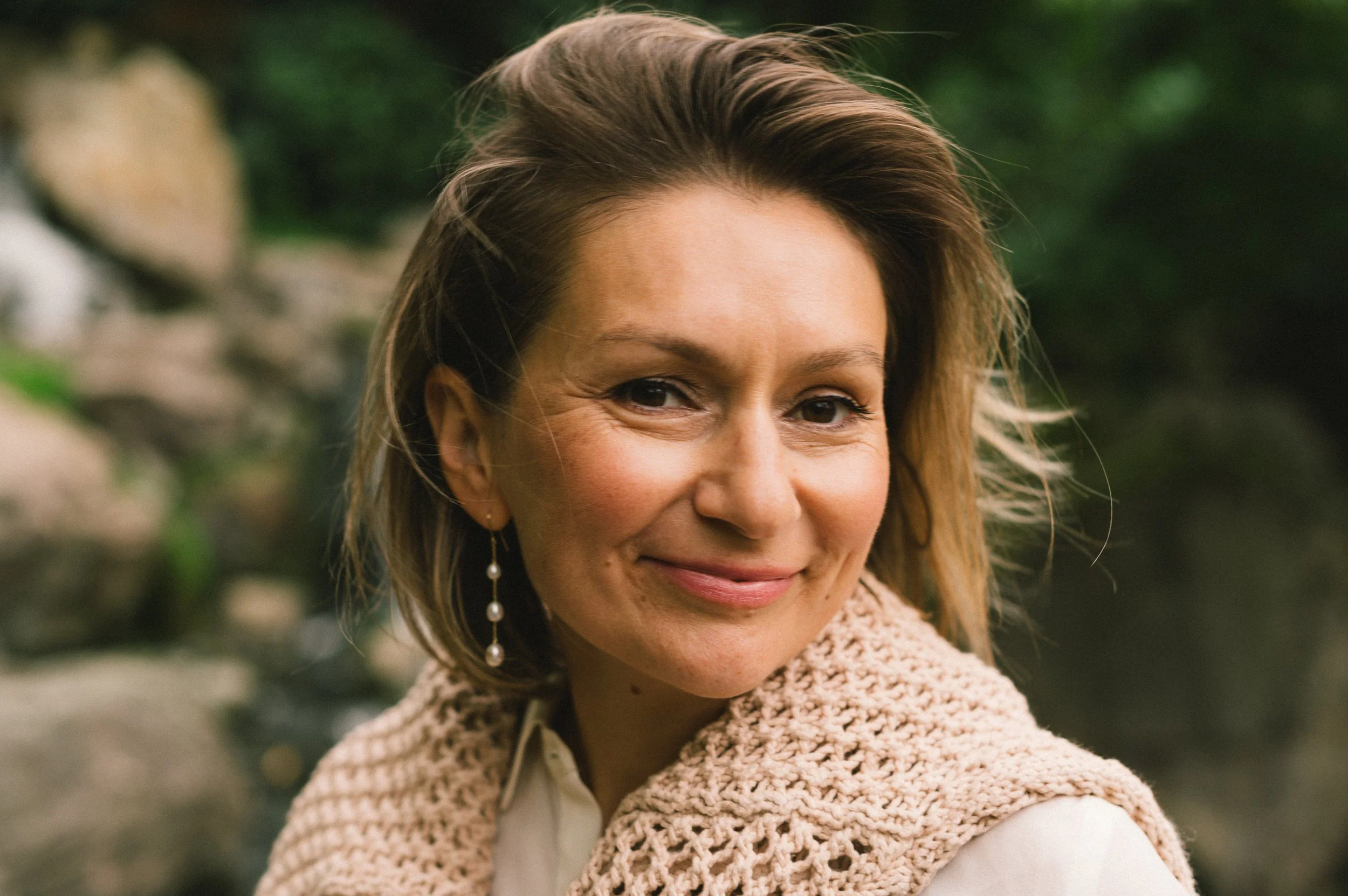 Close-up of a smiling woman with light brown hair, earrings, and a beige knitted shawl outdoors with blurred greenery background.