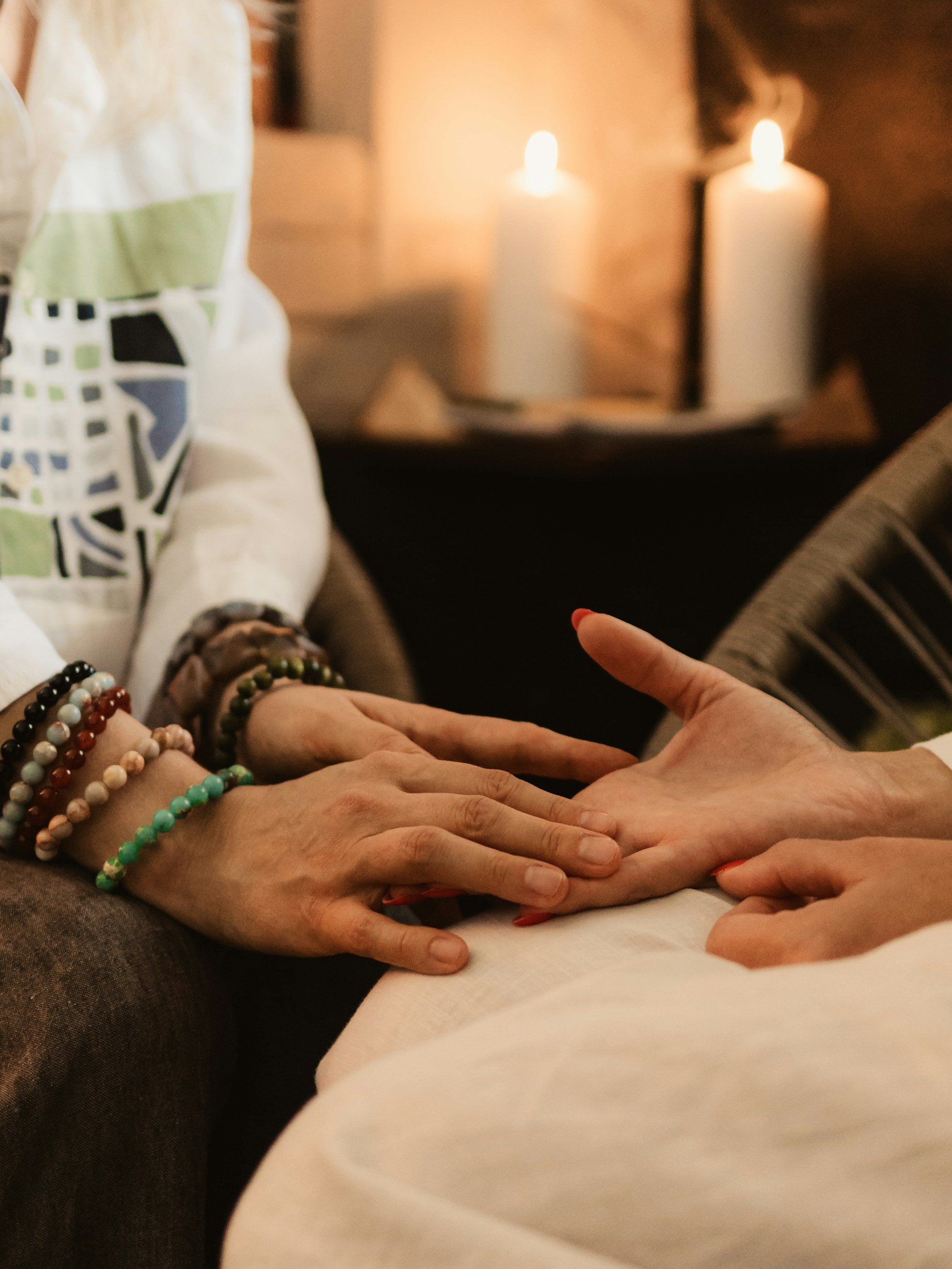 Close-up of multiple hands layered on top of each other in a healing or comforting gesture, with beaded bracelets on one wrist, in a warm indoor setting with candles in the background.