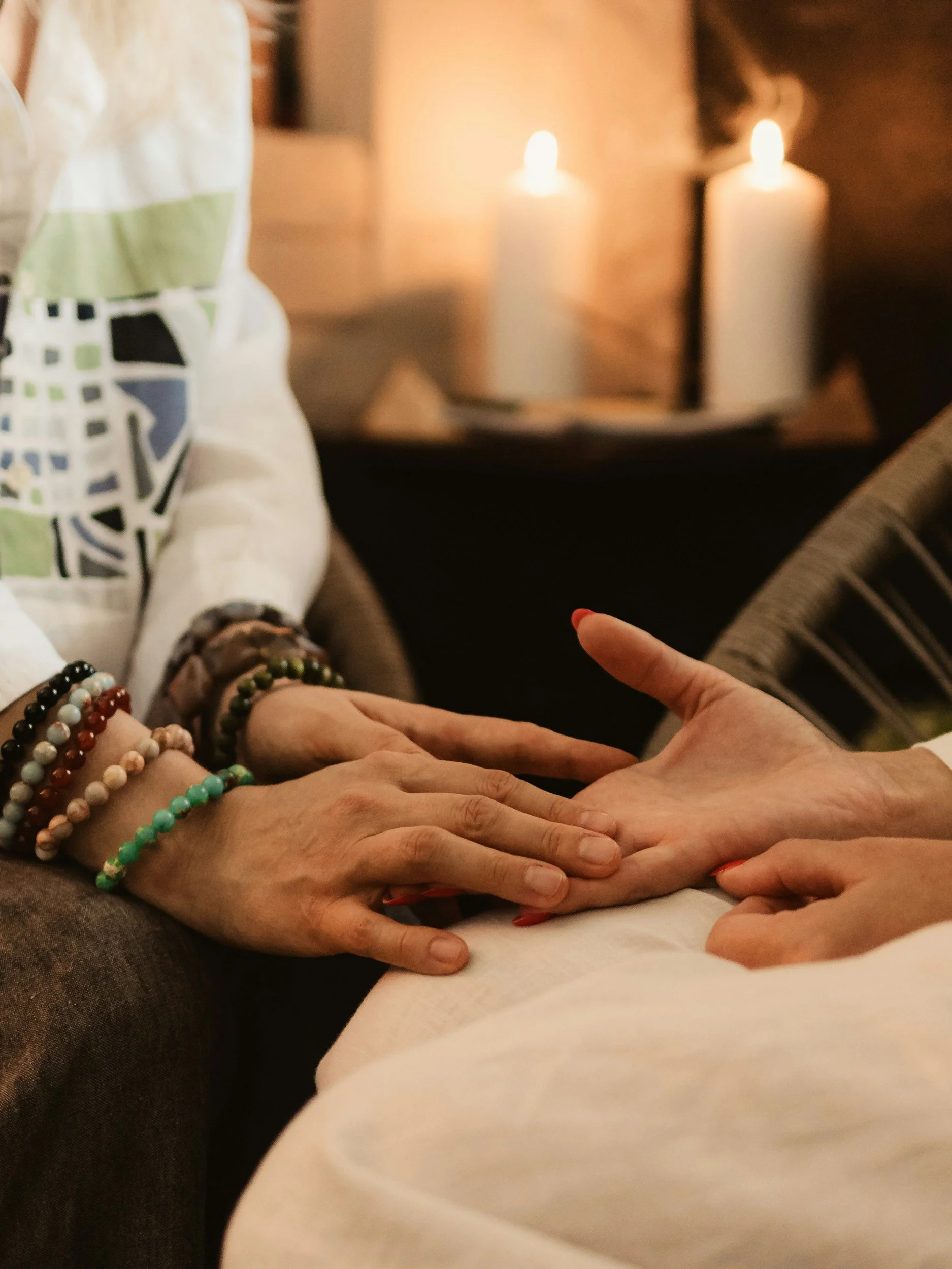 A close-up of two pairs of hands gently touching each other, with a background of lit candles. One hand is adorned with colorful beaded bracelets.
