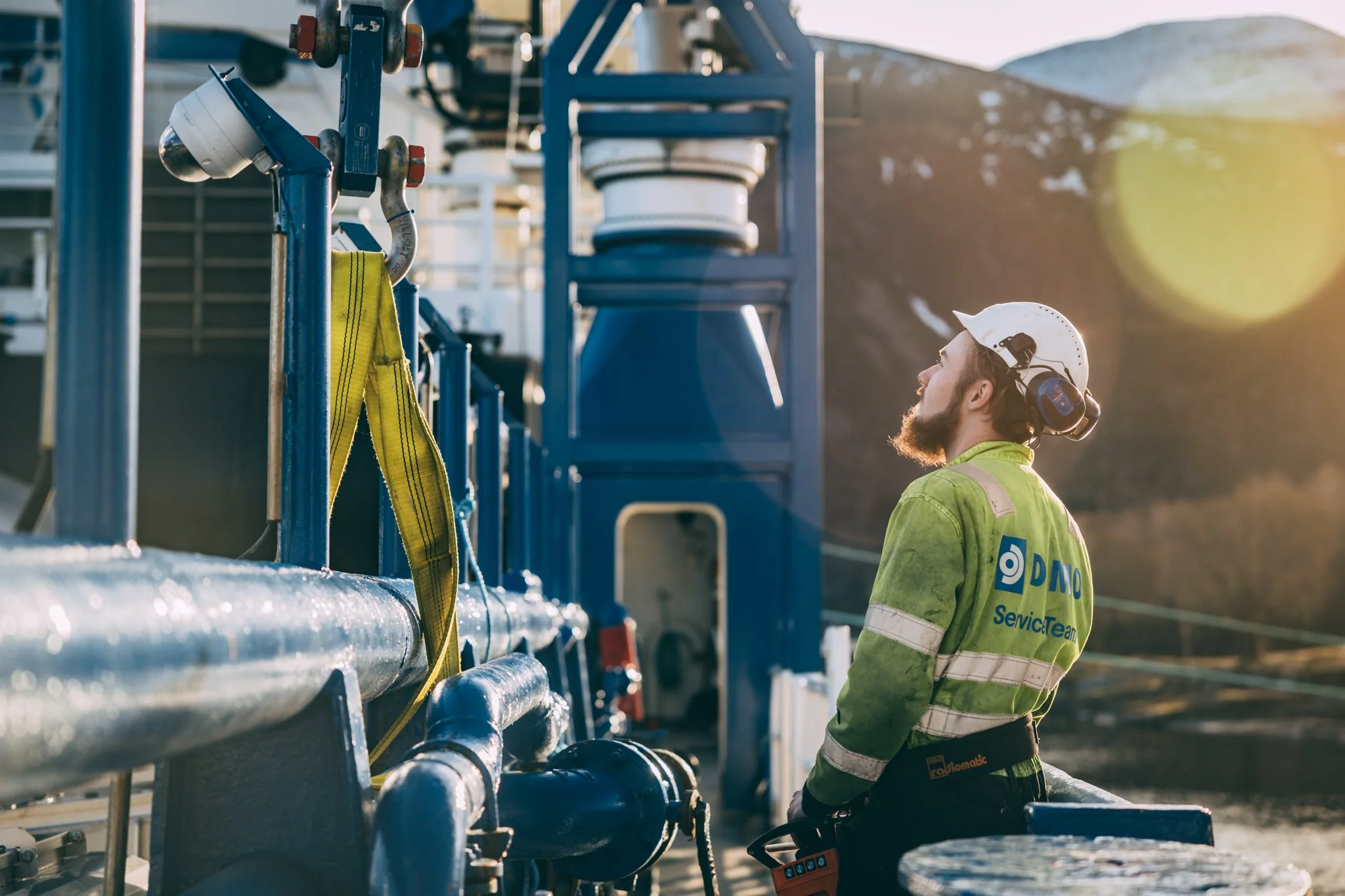 Dimo tar krankontroll på Frøy Challanger, FroyChallanger. Thomas i hørselsvern og refleksvest jobber med industriell kran, med fjell i bakgrunnen og solnedgang. Kran HS Marine.