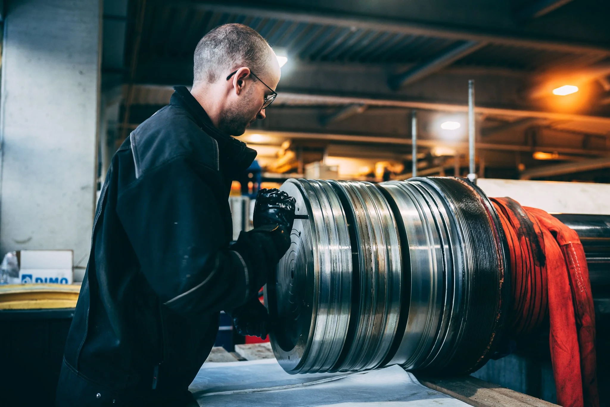 En mann med briller og svart jakke jobber med store metalldeler i et industrilokale.
