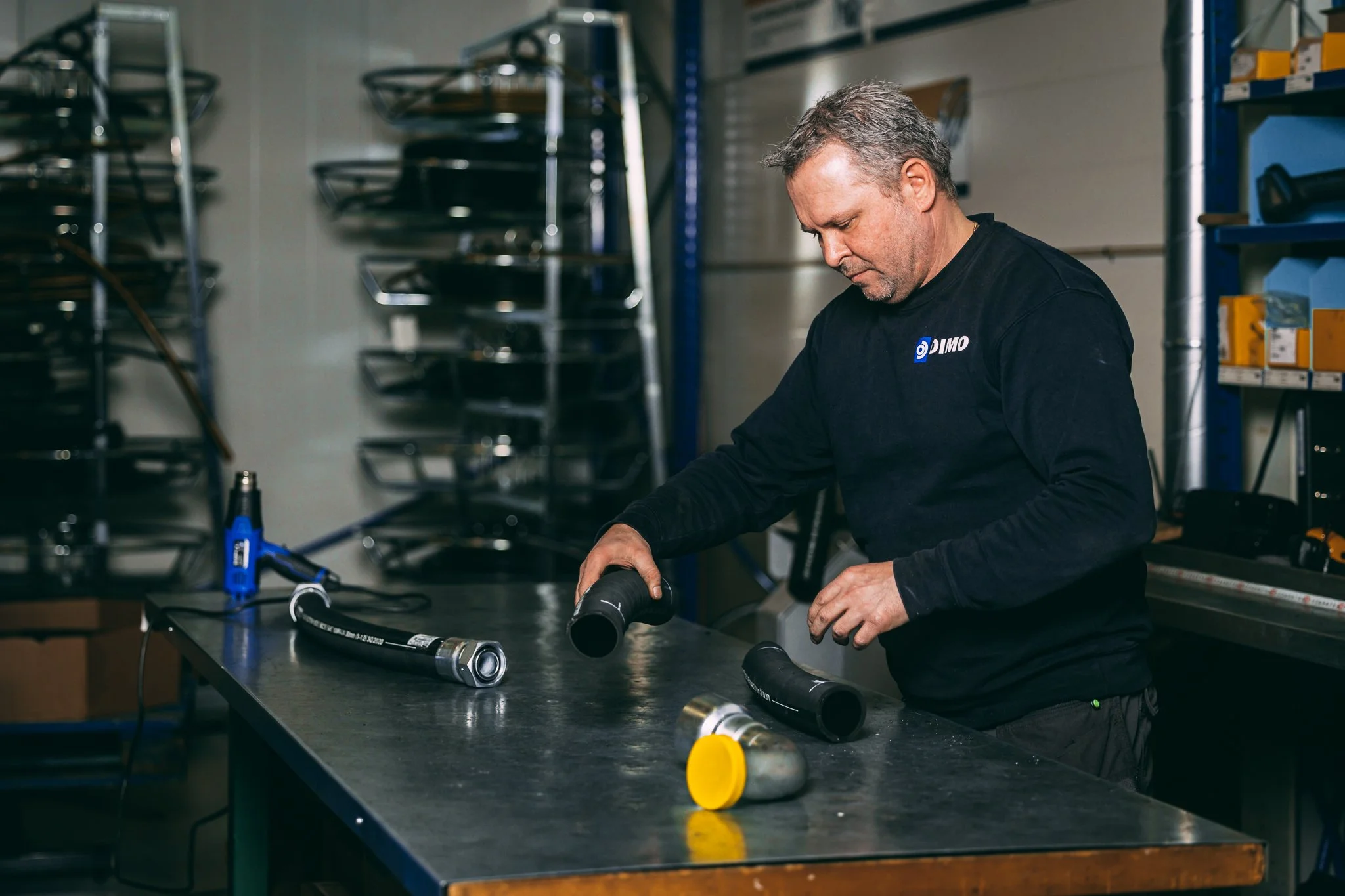 Parker Store, Hose Doctor. En mann arbeider på et verkstedbord, slangeverkstad, omgitt av utstyr og hyller med deler. Breivika, Breivik, Ålesund. Dimo.